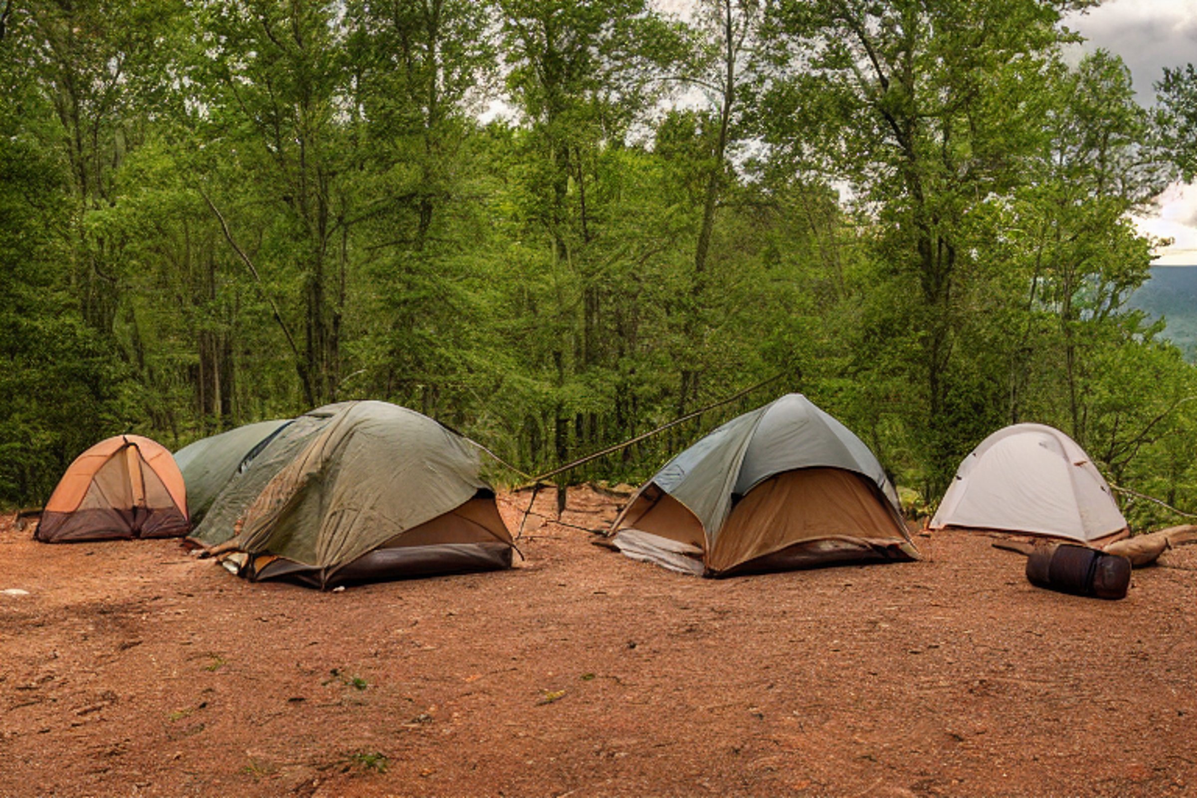 A brown tent rests on a dirt clearing in the Ozark woods. Camp chairs circle a small fire, with a wooden guitar lying nearby beside a large rock. Stormy skies give the campsite a cool, wild feeling.