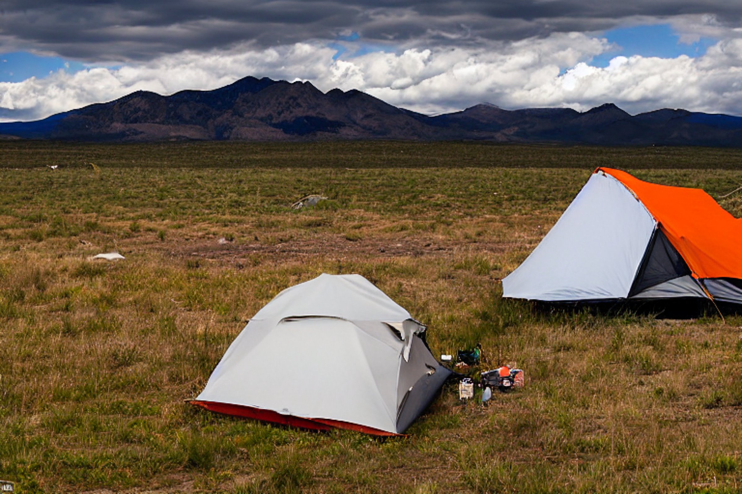 A black tent rests on a dirt patch at a quiet campsite in the Idaho plains. Mountains rise in the distance beneath a cloudy night sky. Camping gear and a clothesline add a cozy, lived-in feel.