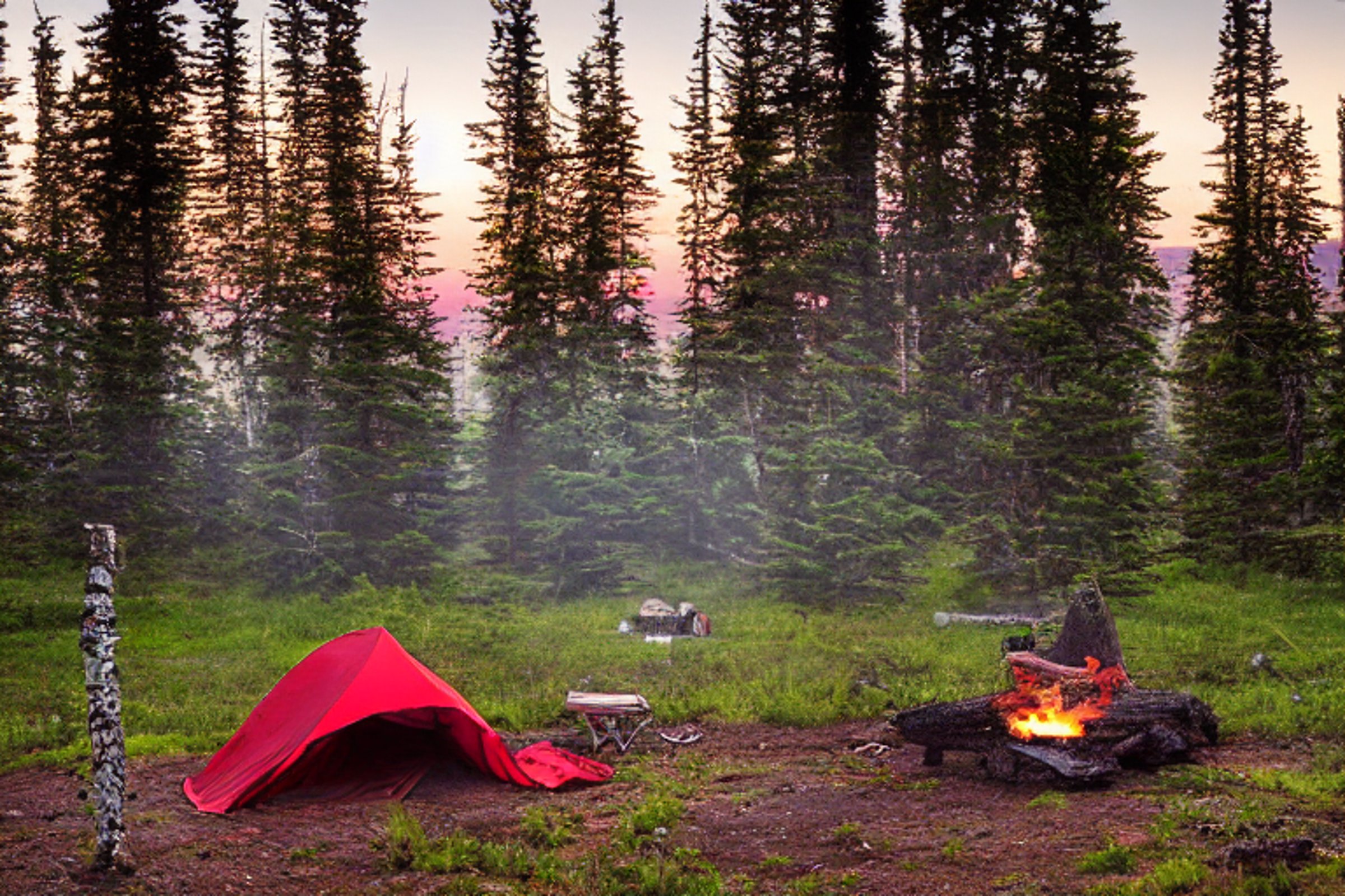 A red tent rests on a quiet dirt campsite in the Alaskan wilderness. Boulders, stones, and a hammock surround a warm campfire as fog drifts through the trees. The sunset gives the peaceful scene a soft, golden glow.