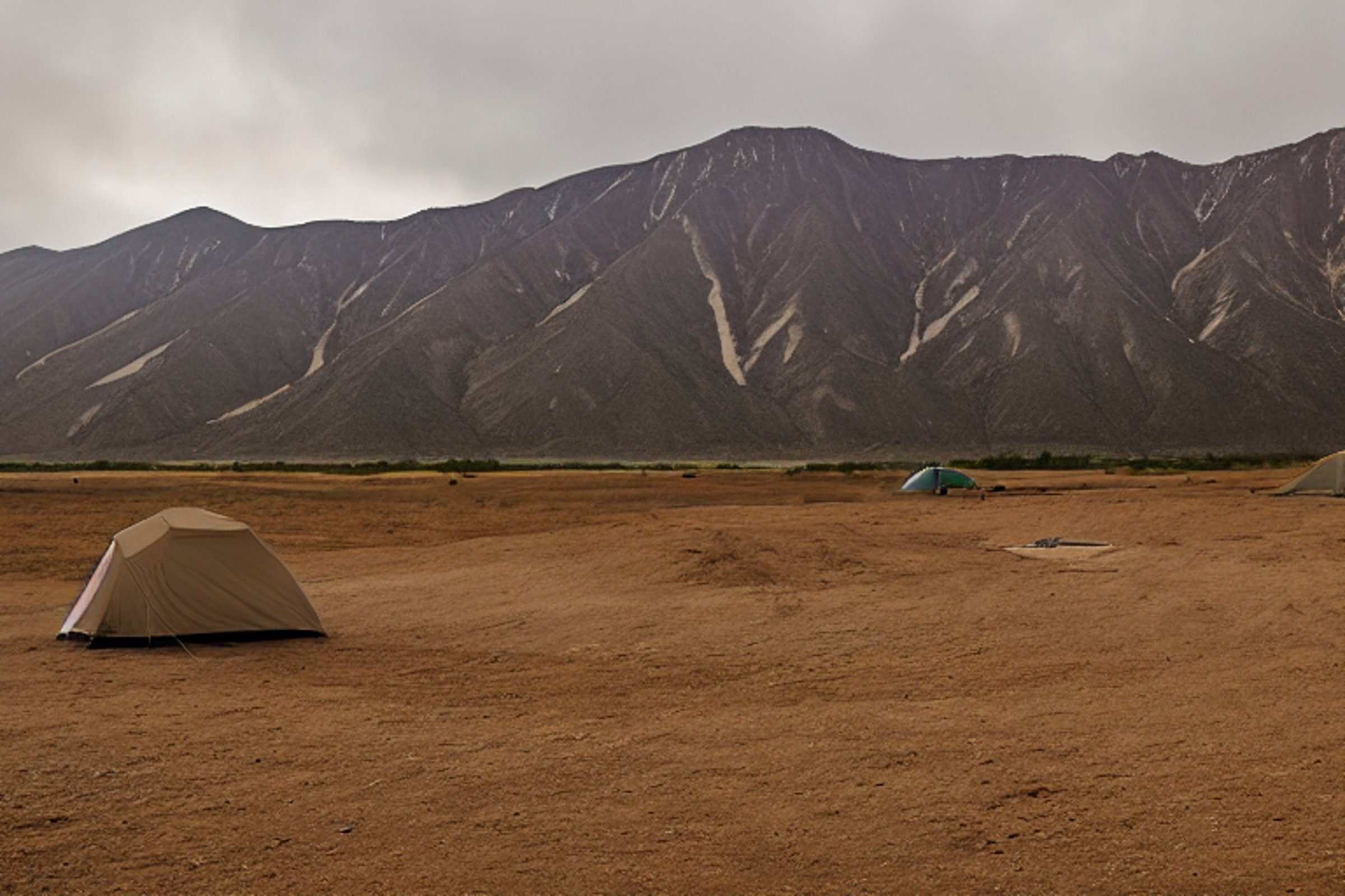 A tan tent rests on a quiet dirt campsite beneath the foggy sky. Hikers explore nearby, while large stone slabs and a parking spot add to the rugged park setting.