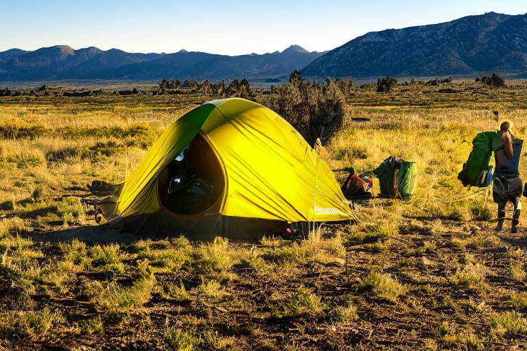 A green tent rests on a sunny paved campsite in the wide Idaho plains. Sunrise lights the distant mountain, while a backpack sits by the dirt patch and a person shines a flashlight nearby.