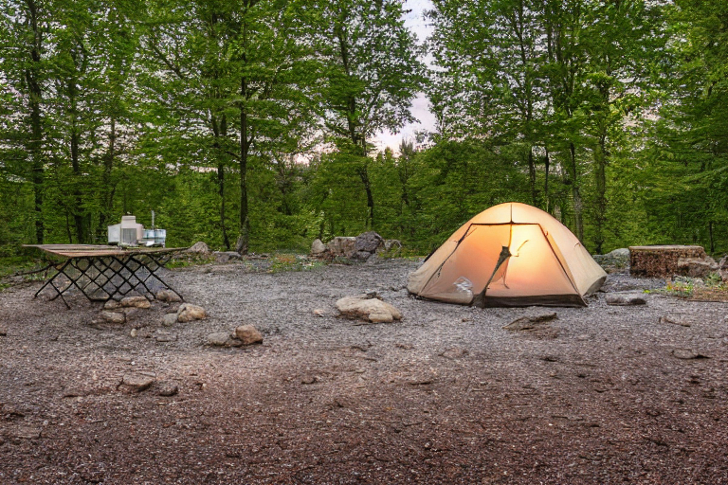 A cozy tent rests beside a paved campsite under a star-filled Upstate New York sky. A warm lantern glows near the gravel and stones, while a picnic table sits nearby.