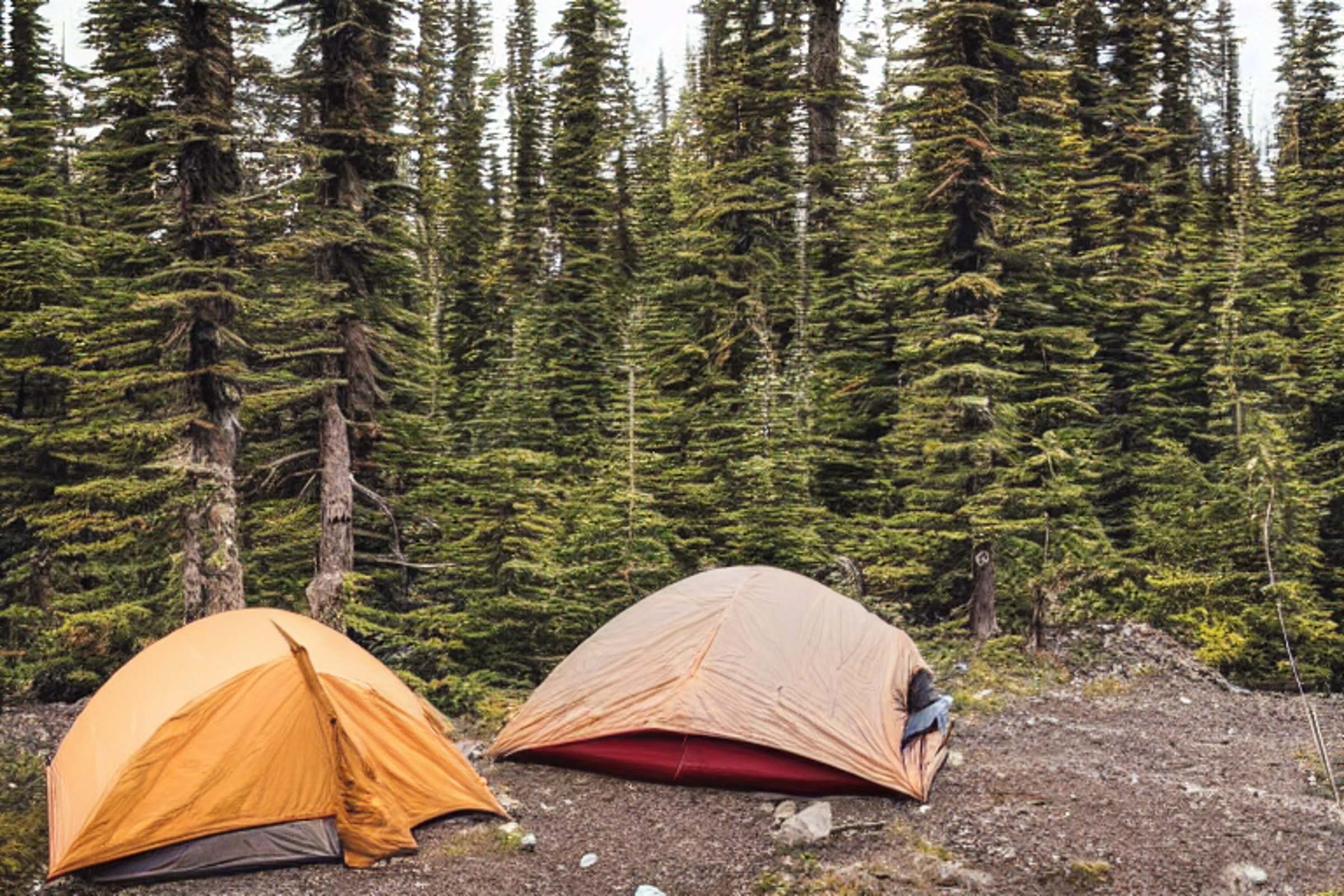 A cozy tan tent rests in a snowy Alaska wilderness beneath a sky full of stars. A warm campfire glows on the rocky ground, with a backpack nearby and clothes hanging on a line.