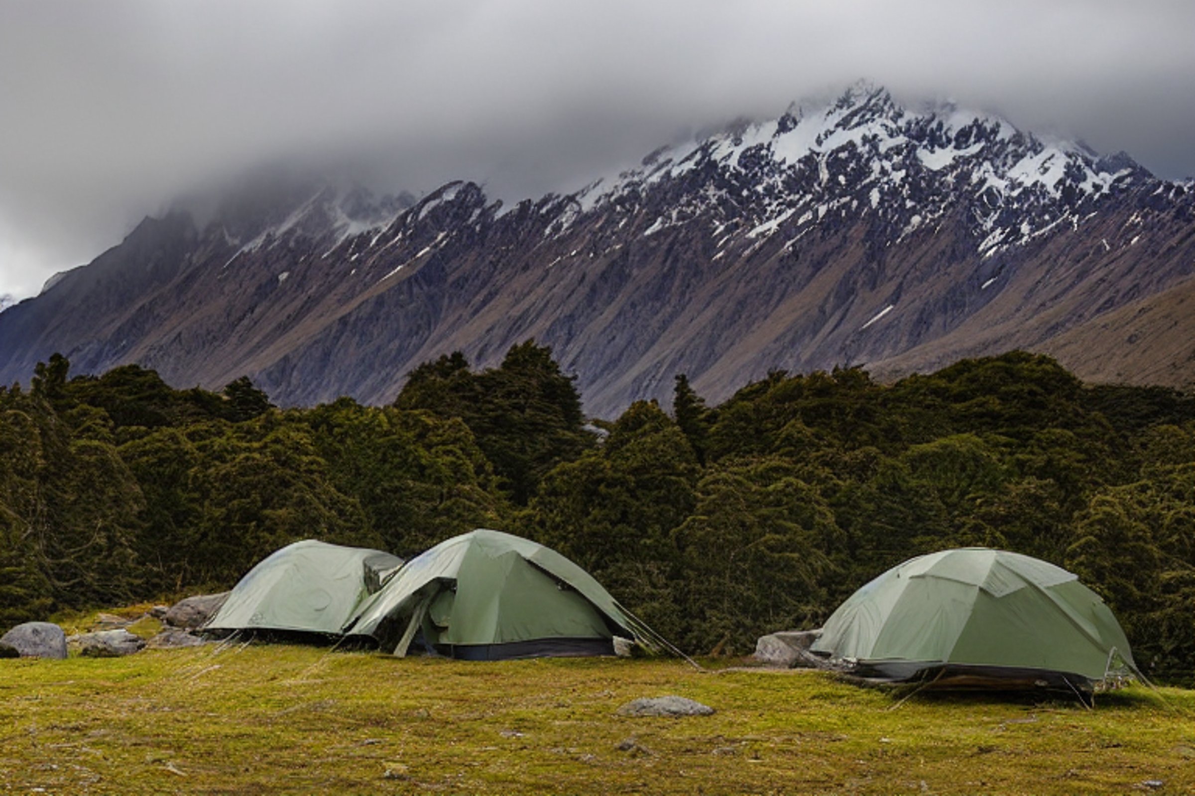 A green tent rests on a dirt clearing beneath the dark, stormy sky of Mount Cook National Park. A warm campfire glows beside stone slabs and camping chairs, with rolling hills fading into the night.