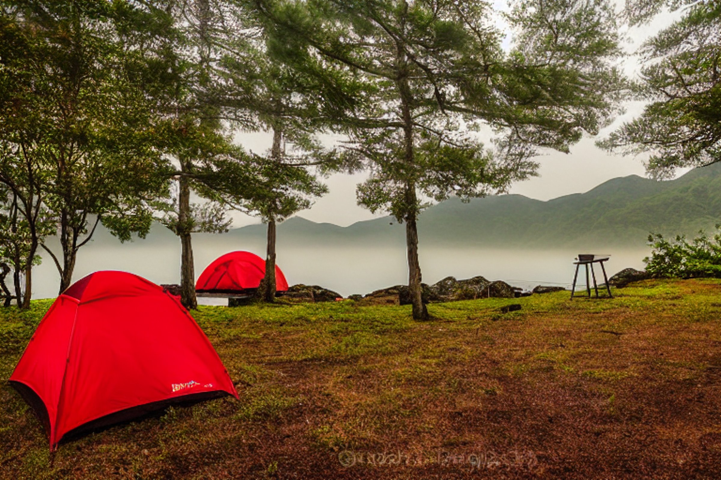 A bright red tent glows beside a small campfire in the misty hills of Miyajima, Japan. Tall grass surrounds the campsite, with a picnic table and a mountain rising in the soft sunrise.