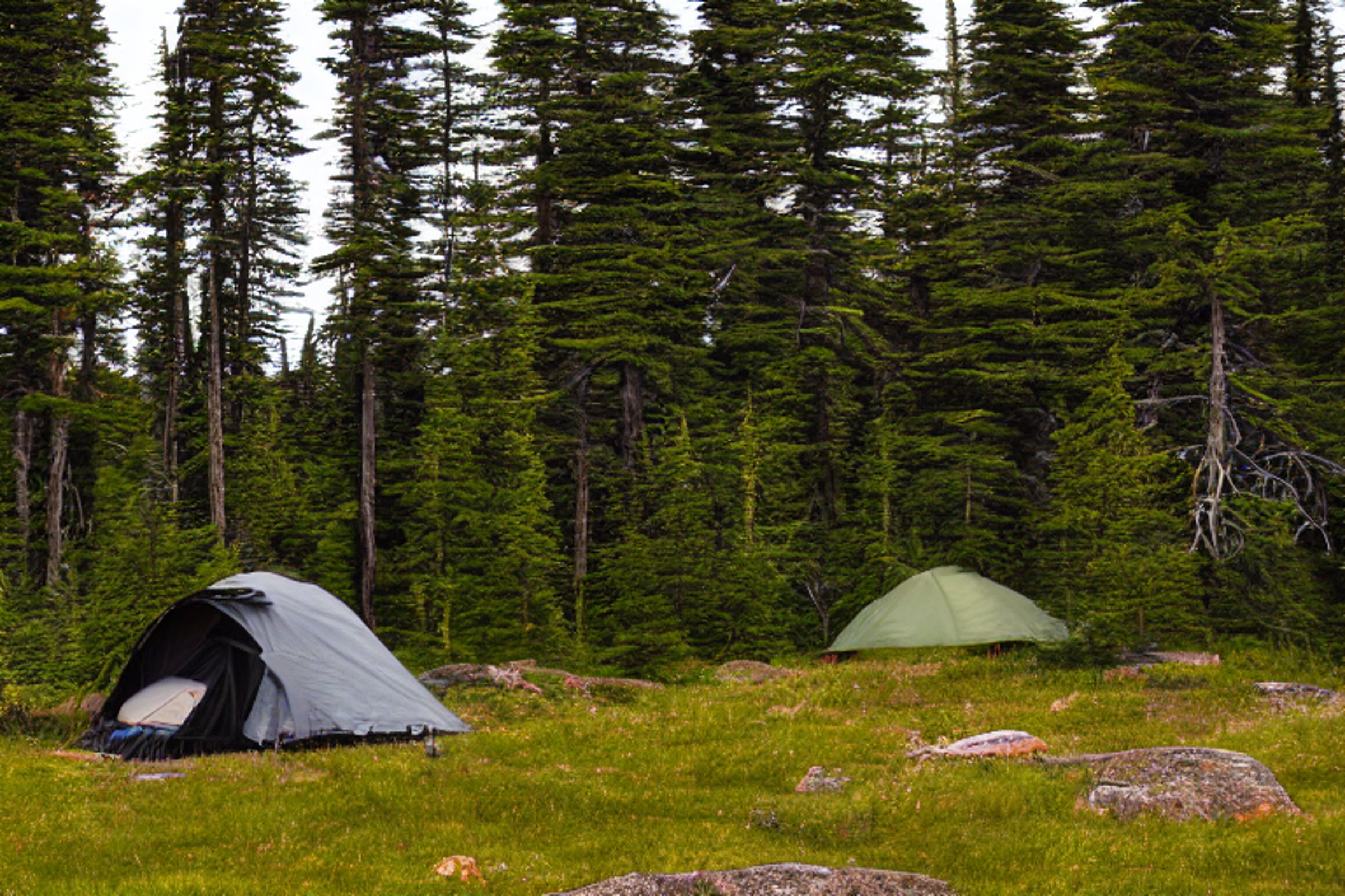 A black tent rests on a paved campsite as soft rain falls at dusk. Rolling hills, large boulders, and a distant mountain surround the site, while a small flashlight glows in the distance.