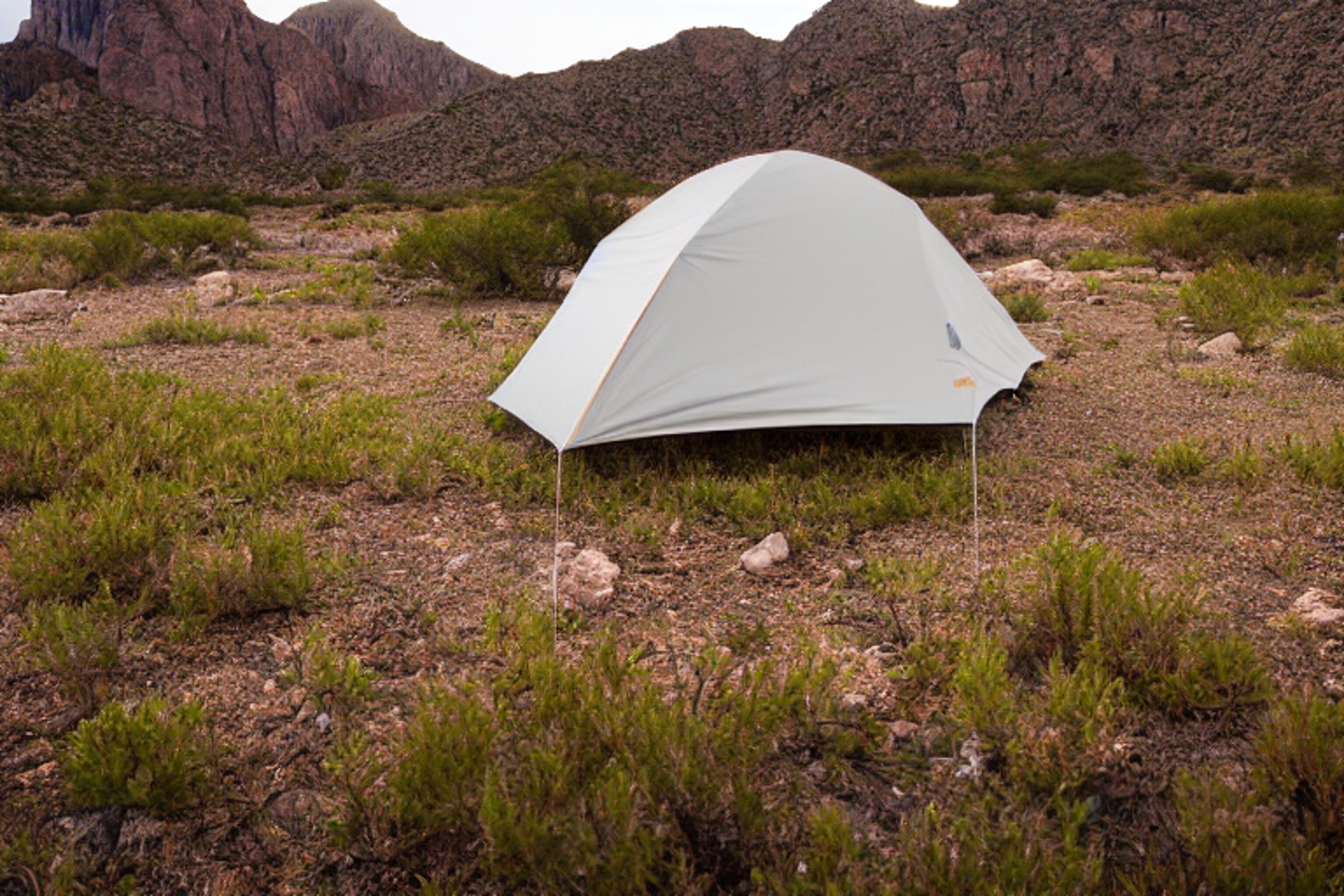 A cozy tent glows softly on a paved campsite in Big Bend National Park. Large stone slabs, fallen leaves, and small flowers surround the dirt clearing. Misty night air gives the camp a quiet, peaceful feel.