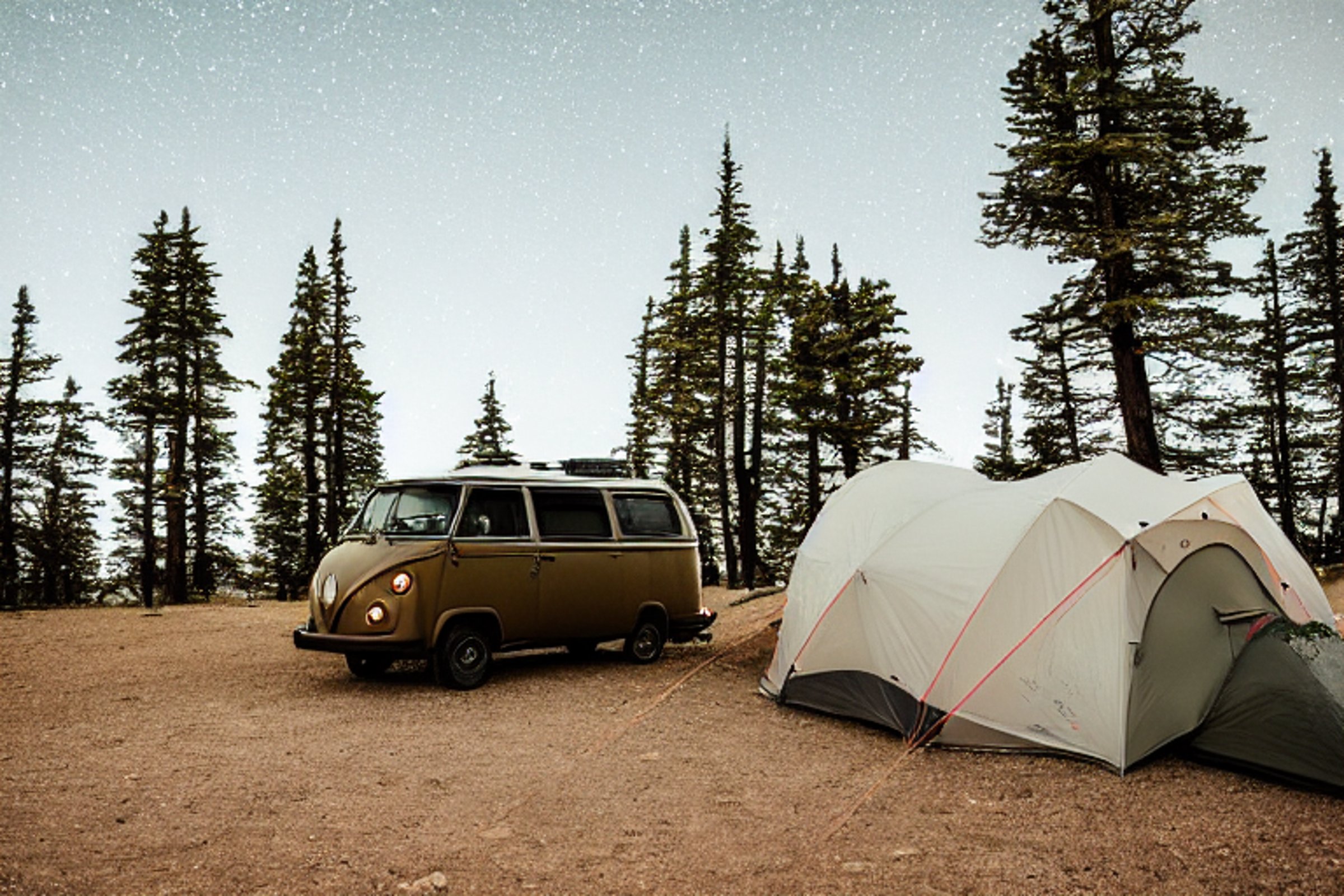 A black camper van rests beside a warm campfire under glowing northern lights. Fog drifts around the sandy dunes, small trees, and stone-covered ground, while a nearby tent shines with soft light.