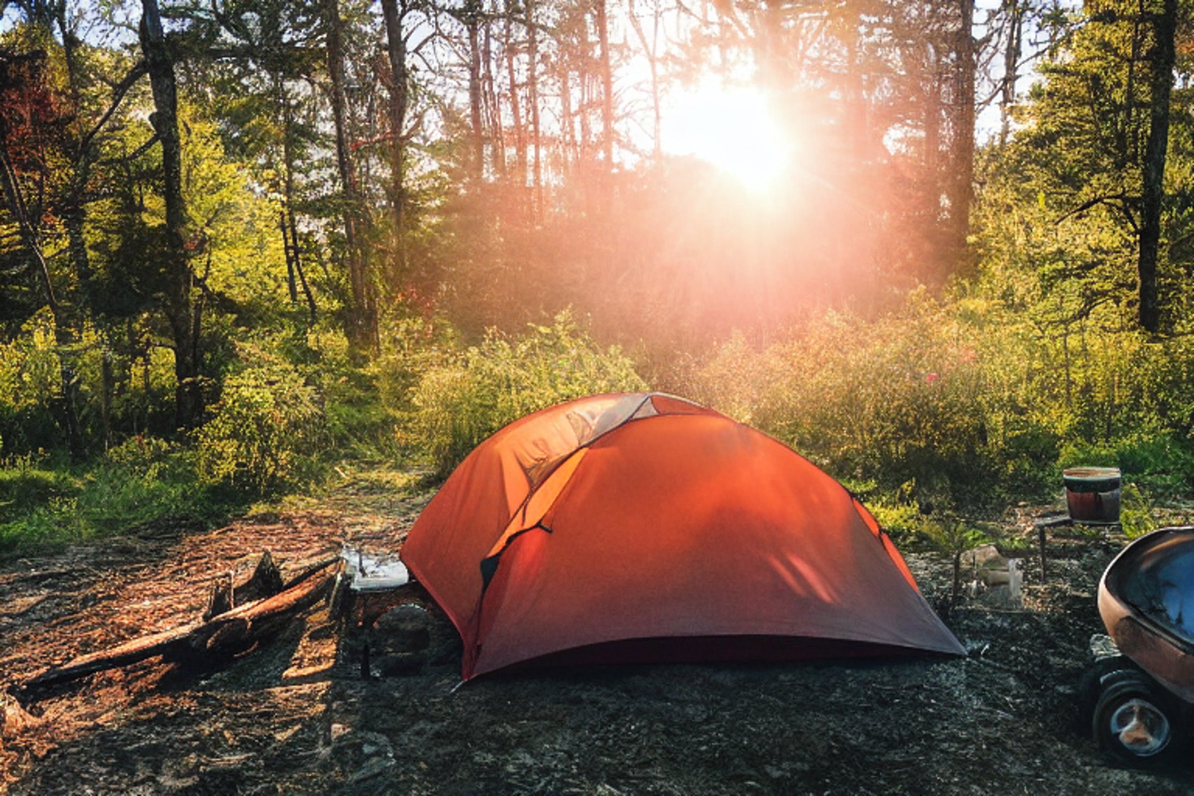 A colorful tent rests on a quiet dirt campsite in the Smoky Mountains. A warm campfire glows beside the logs as rolling hills stretch into the sunset. Sunlight fills the peaceful scene.