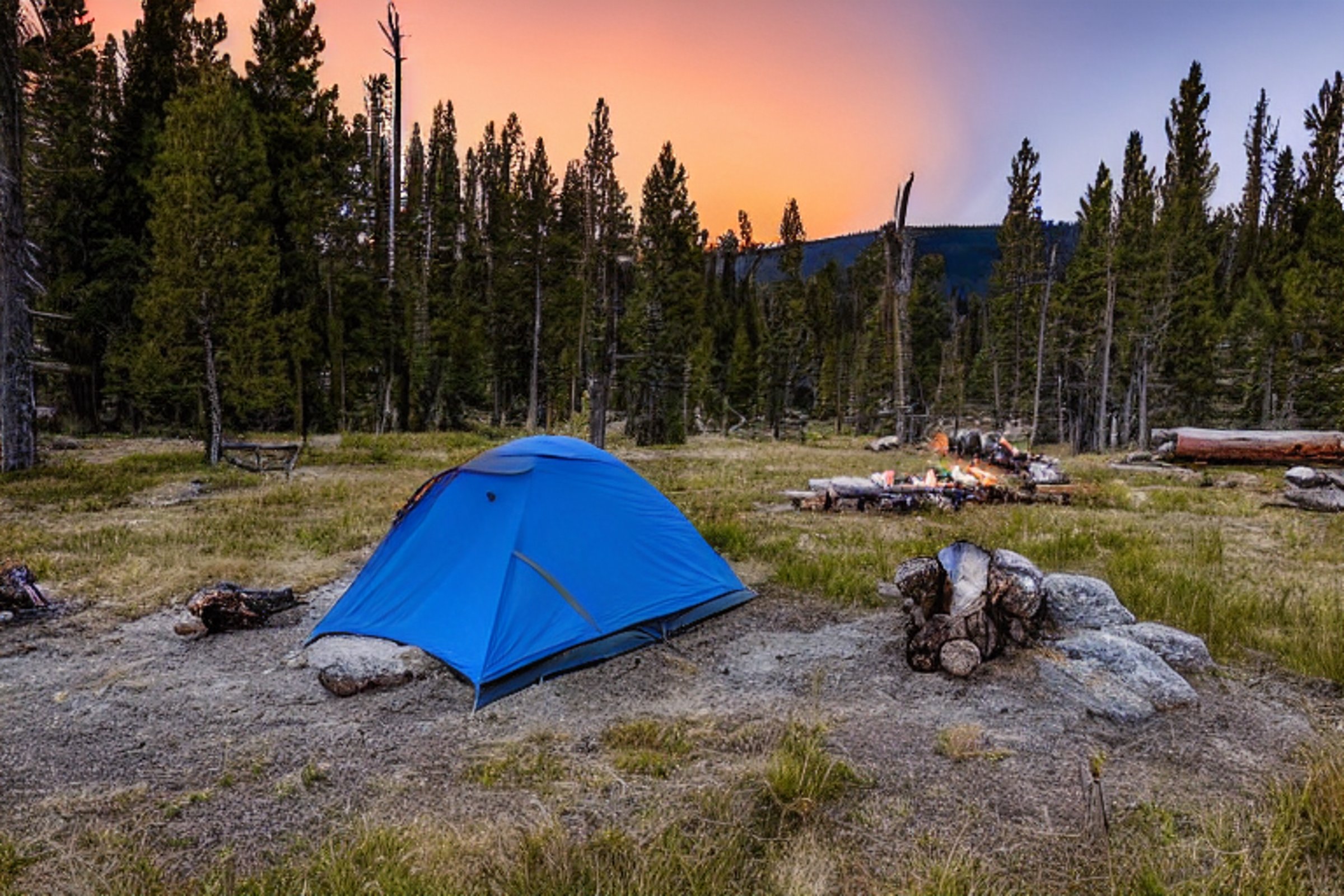 A gray and blue tent rests on a dirt clearing in Yellowstone as the sun sets. A warm campfire glows beside a large log and scattered boulders, while hikers enjoy the peaceful evening. A pale moon rises over the wide, sunny landscape.