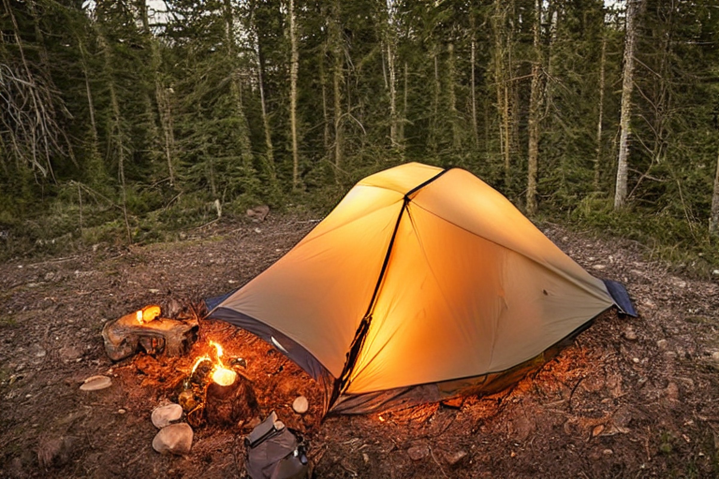 A cozy tan tent rests beside a quiet river as the sun sets. A warm campfire glows on the dirt, with logs, a backpack, and a picnic table nearby. The clear sky and soft light create a peaceful camping scene.