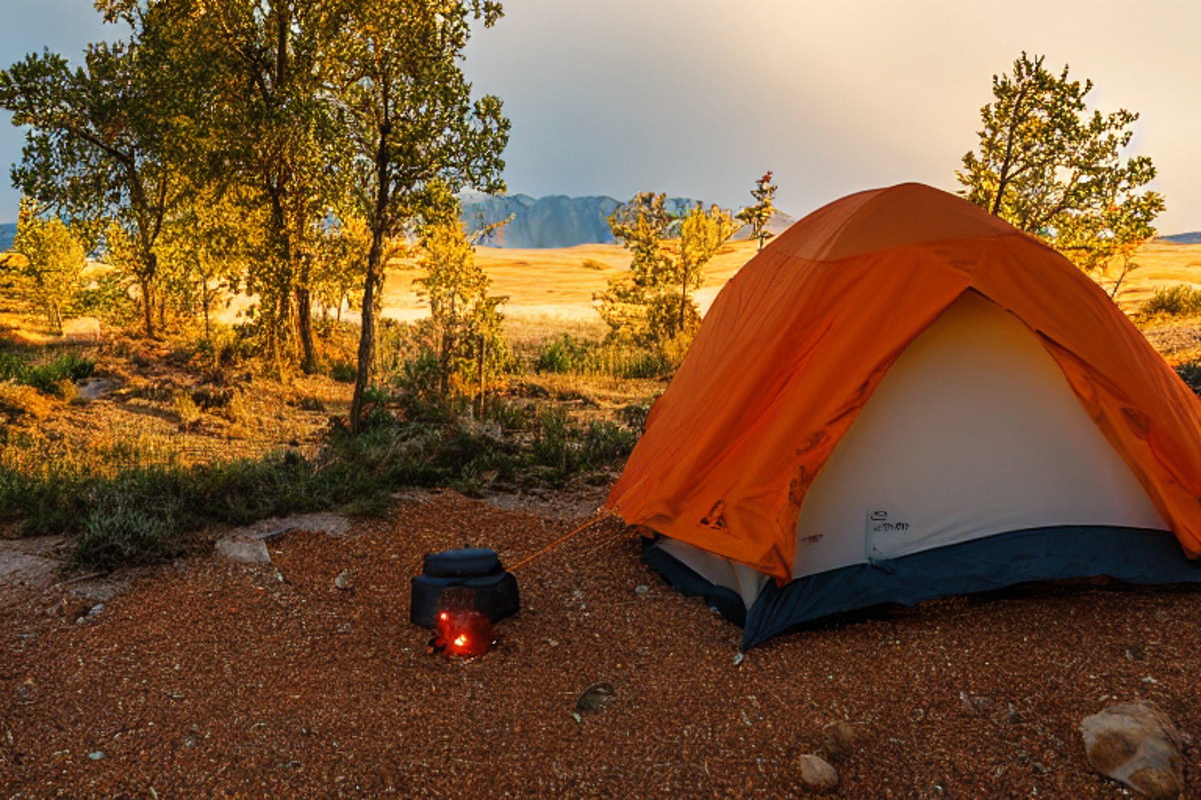 An orange tent rests beneath tall trees beside a glowing campfire. Rainy dusk settles over the dirt campsite, with leaves, a lantern, and a large rock nearby. Beyond the trees, the Great Sand Dunes add a wild and peaceful touch.