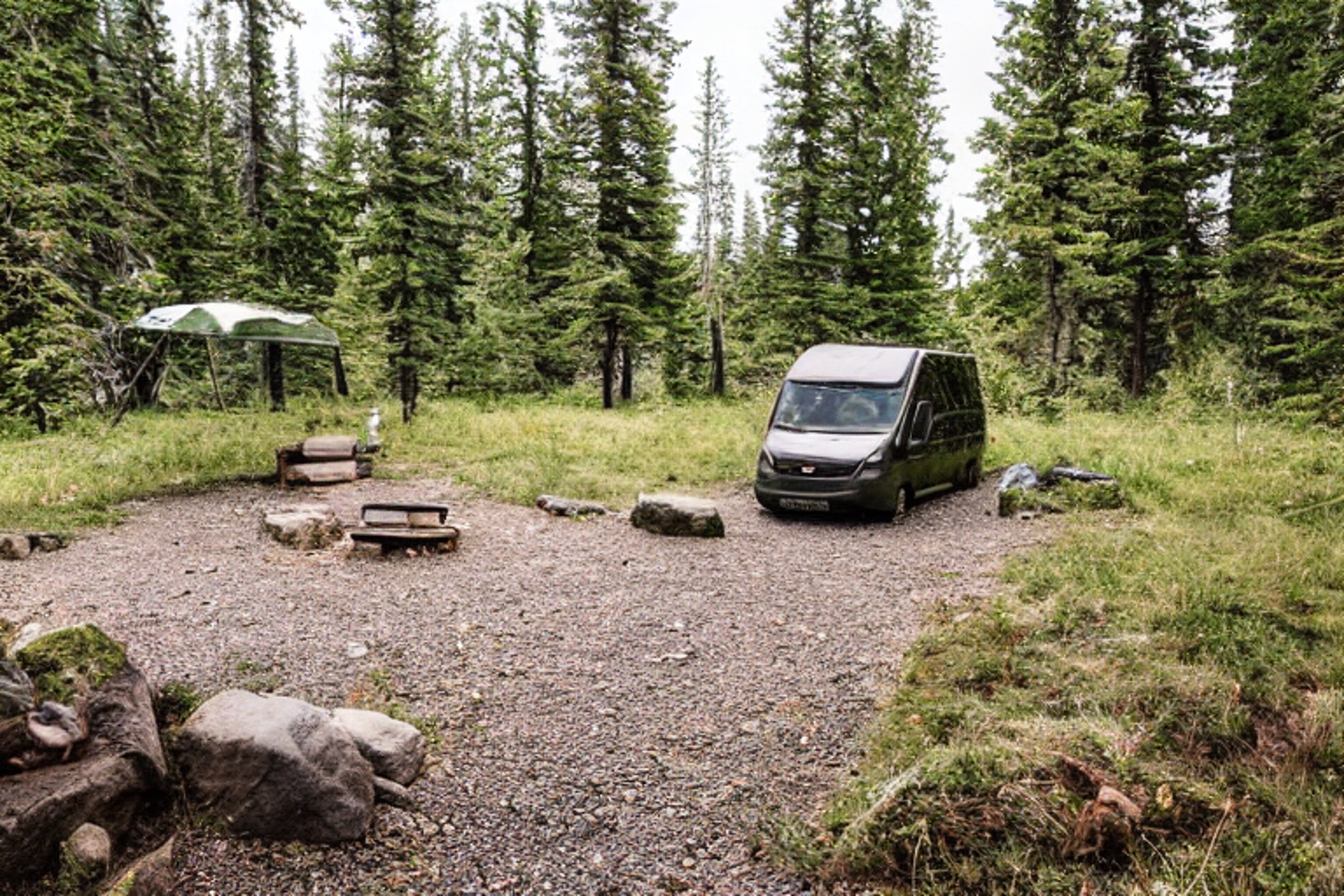 A green camo van rests at a quiet campsite beneath a starry night sky. Rainy weather, rocky ground, and a gentle stream create a peaceful mountain setting. A hammock hangs between the trees nearby.
