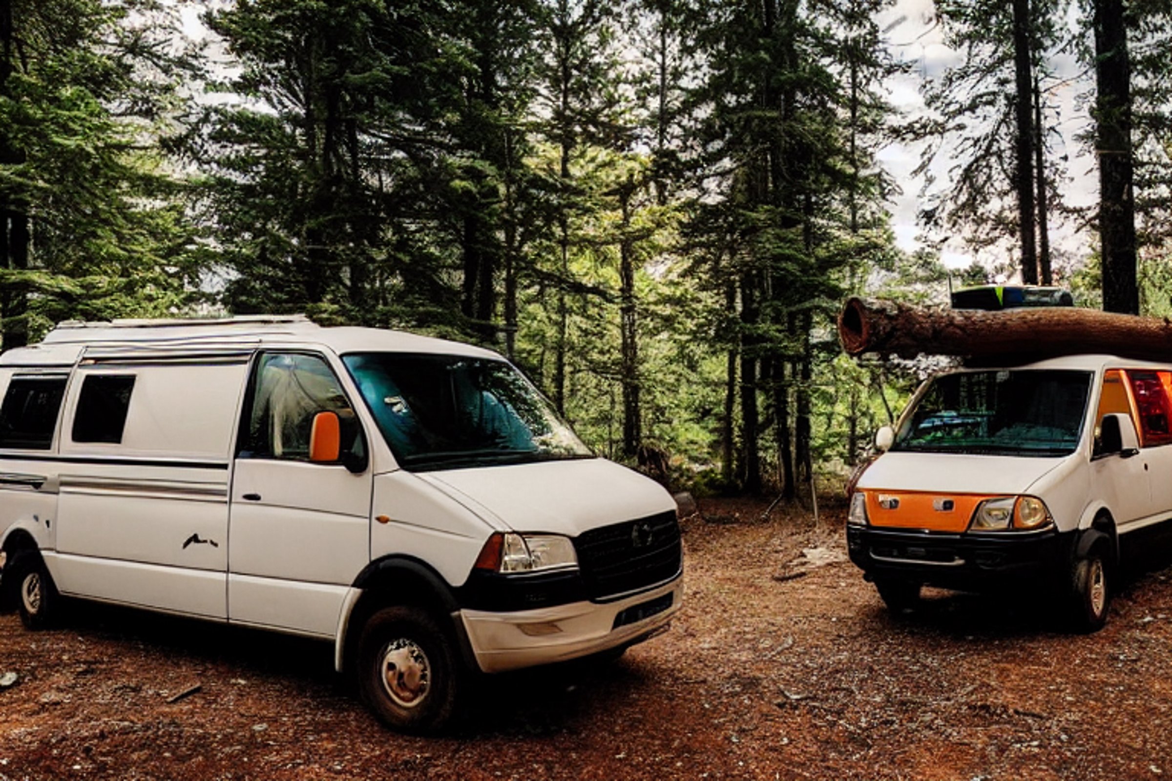 A colorful camper van rests beside a quiet creek in a foggy forest. A warm campfire glows on the dirt, with logs, a picnic table, and a lantern nearby as the sun sets.