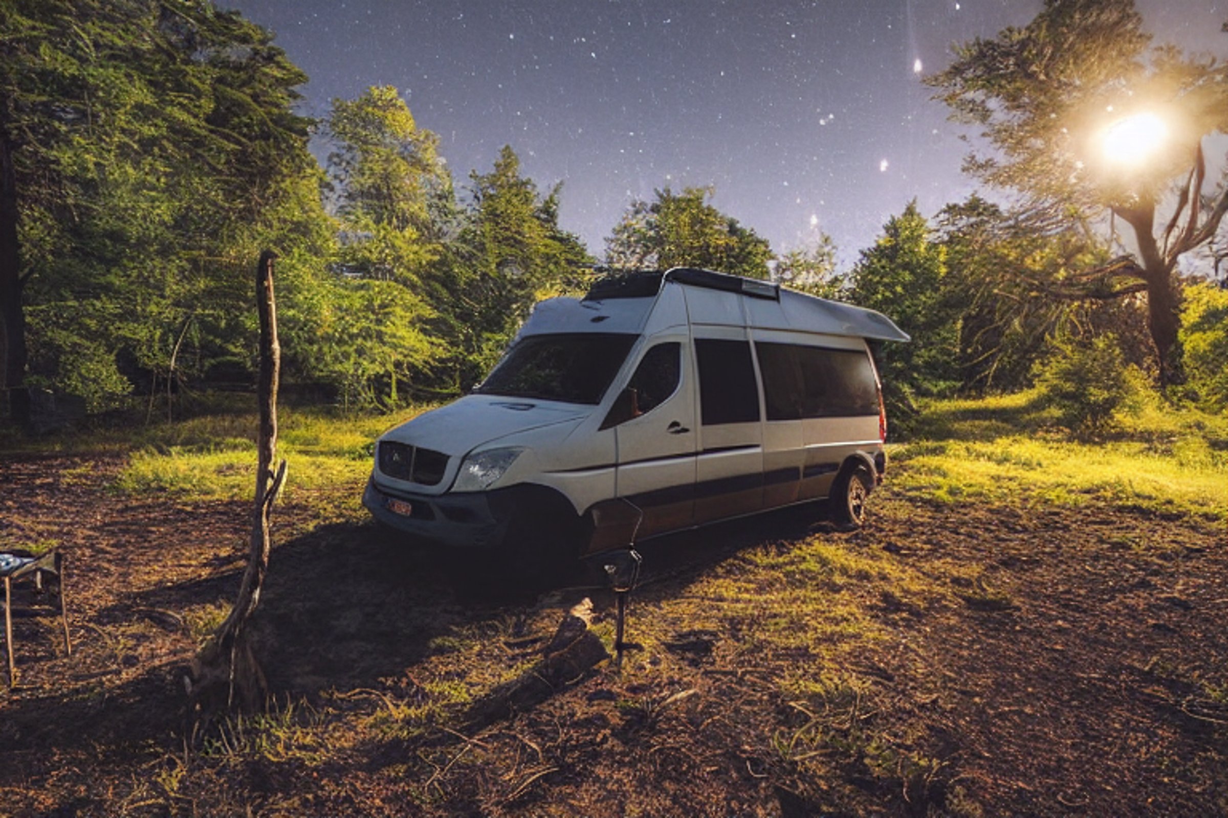 A cozy camper van rests on a sunny dirt clearing near a warm hot spring. A hammock hangs between leafy trees, while a stone-ringed campfire glows beside a softly lit tent.
