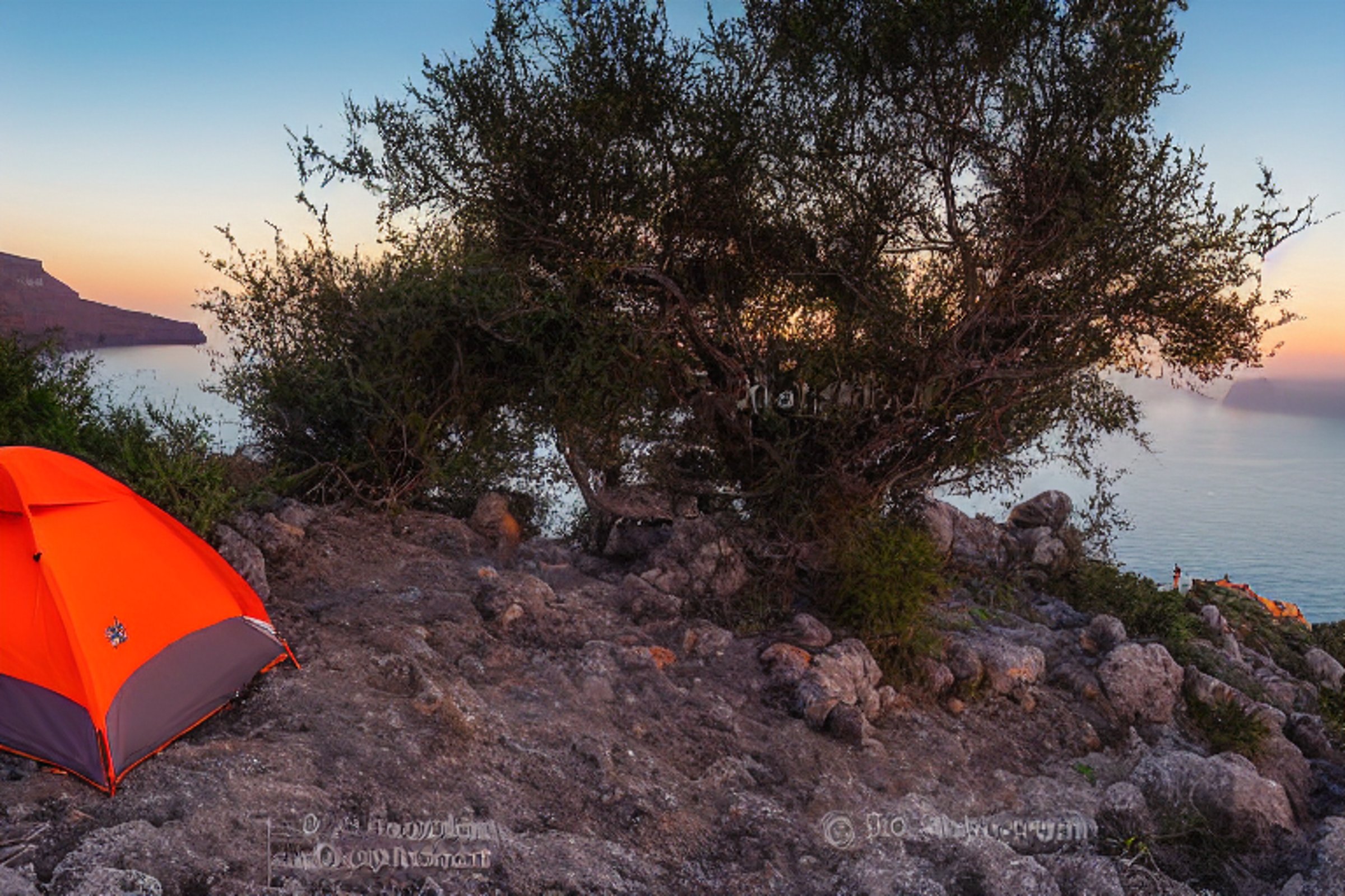 An orange tent rests on a paved campsite in Santorini as the sun sets behind soft clouds. Small trees and boulders surround a dirt patch, while a backpack sits nearby. In the distance, surfers enjoy the coast.