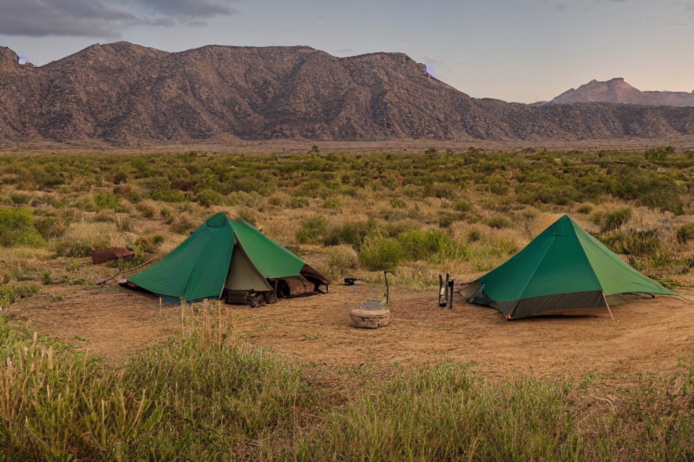 A green and tan tent rests on a dirt campsite in the New Mexico desert. Large bushes surround the camp, with a mountain rising in the distance under a rainy sunset sky. A wooden guitar lies nearby, adding a cozy touch.
