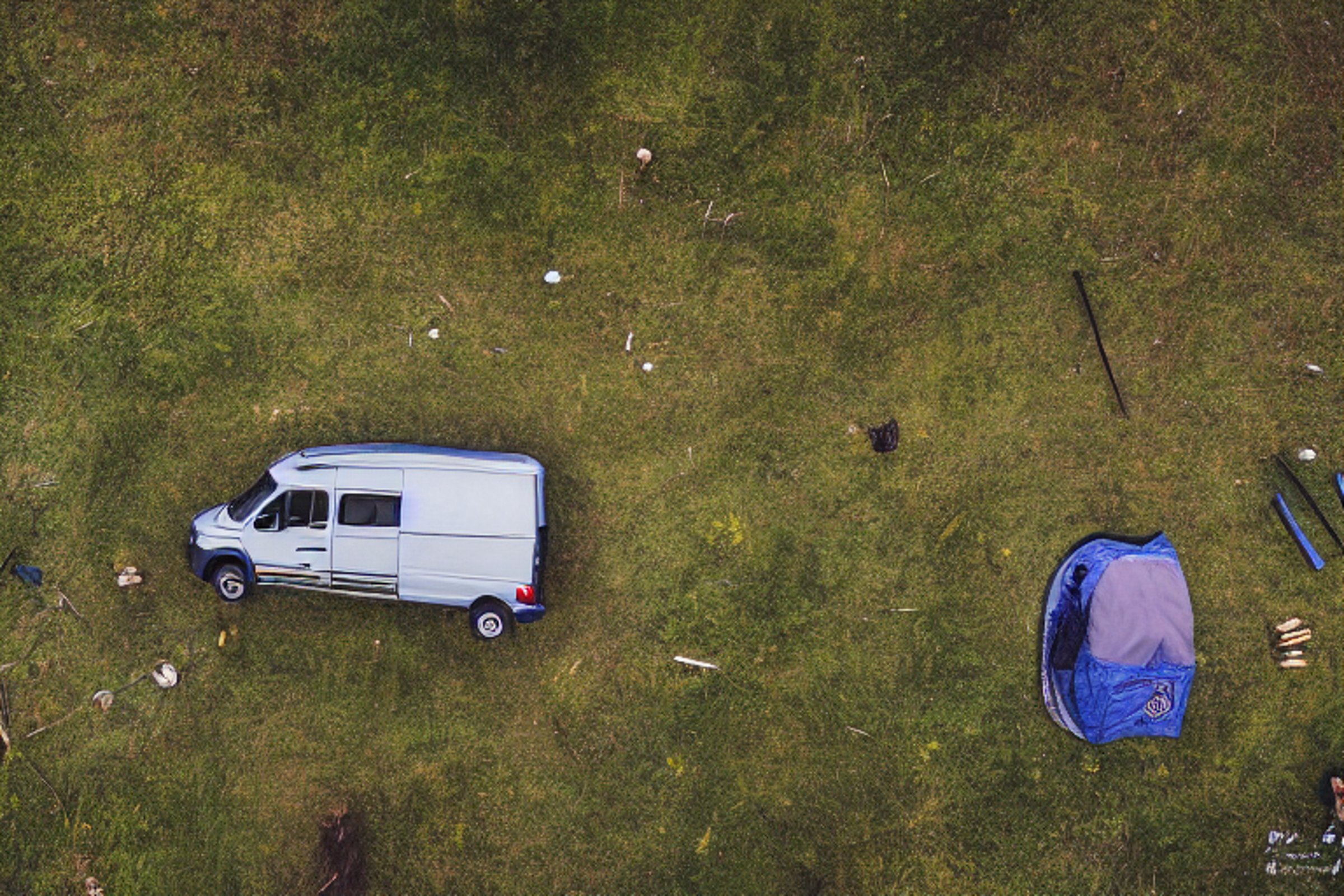 A blue camper van rests in a quiet Alaskan wilderness campsite at sunrise. Tall grass, rough ground, and a warm campfire create a cozy feeling under the snowy sky. A glowing tent waits nearby for a night of adventure.
