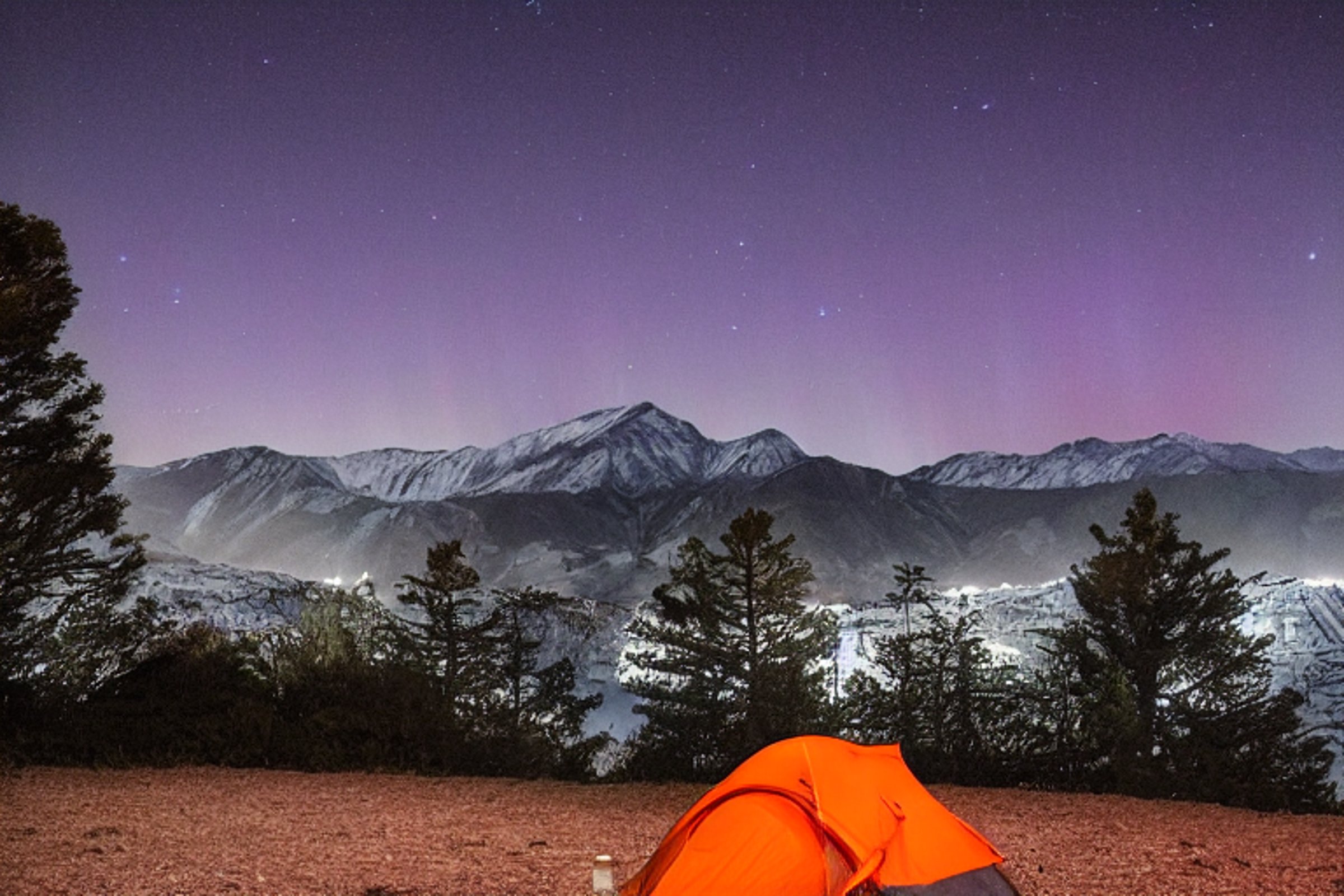 A gray and orange tent glows softly on a quiet campsite beneath the colorful northern lights. Mountains and small trees surround the dirt clearing, while clothes hang from a nearby line.