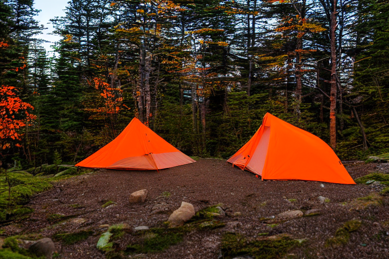 A gray and orange tent glows softly on a dirt campsite as the sun sets in Acadia National Park. Fallen leaves surround the tent and picnic table, creating a warm, peaceful outdoor scene.