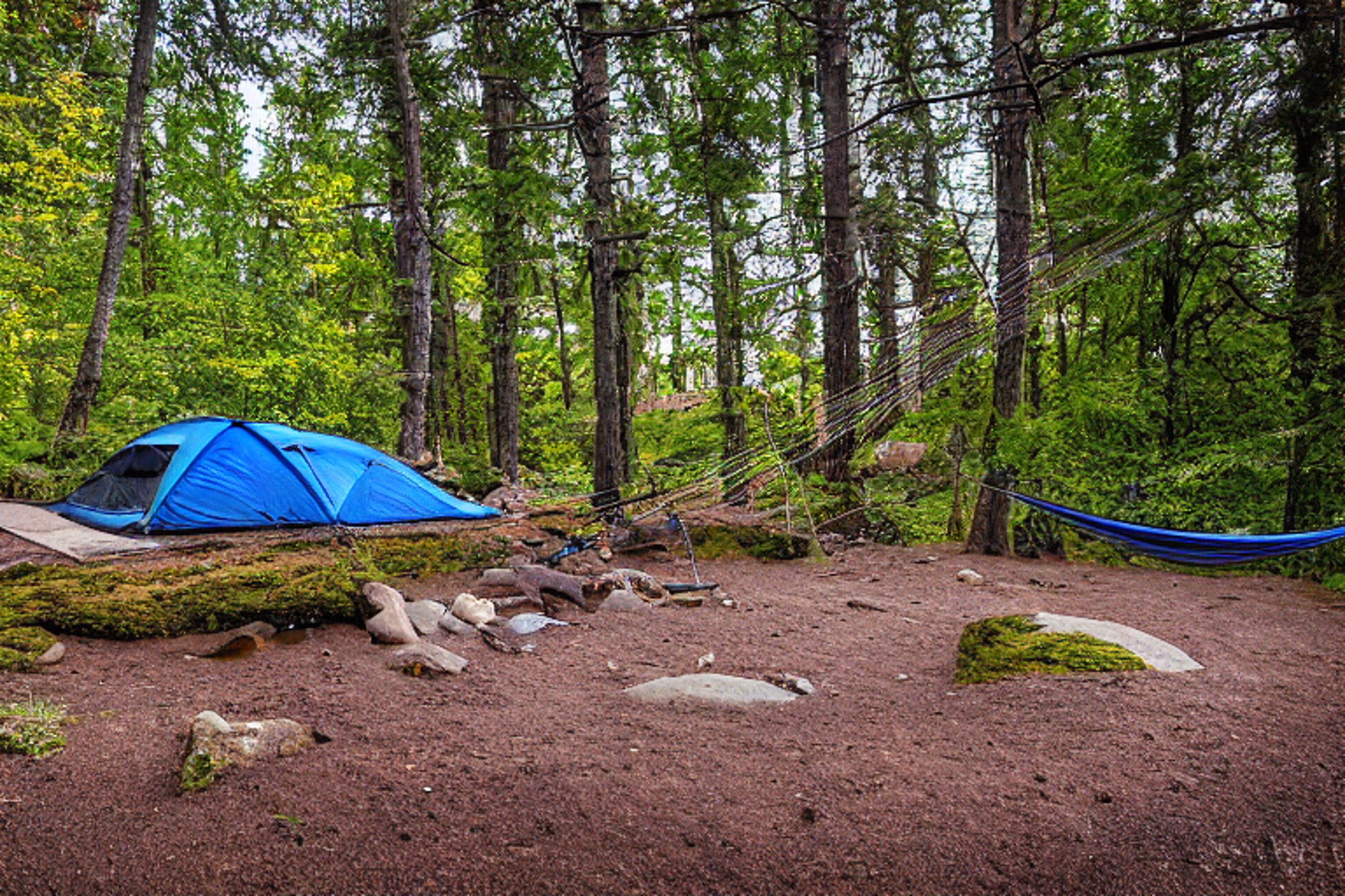 A blue and gray tent rests on a snowy dirt clearing beside a gentle stream. A hammock hangs between the trees, while large boulders add a rugged touch. The soft sunset light creates a peaceful feeling in this North Shore campsite.