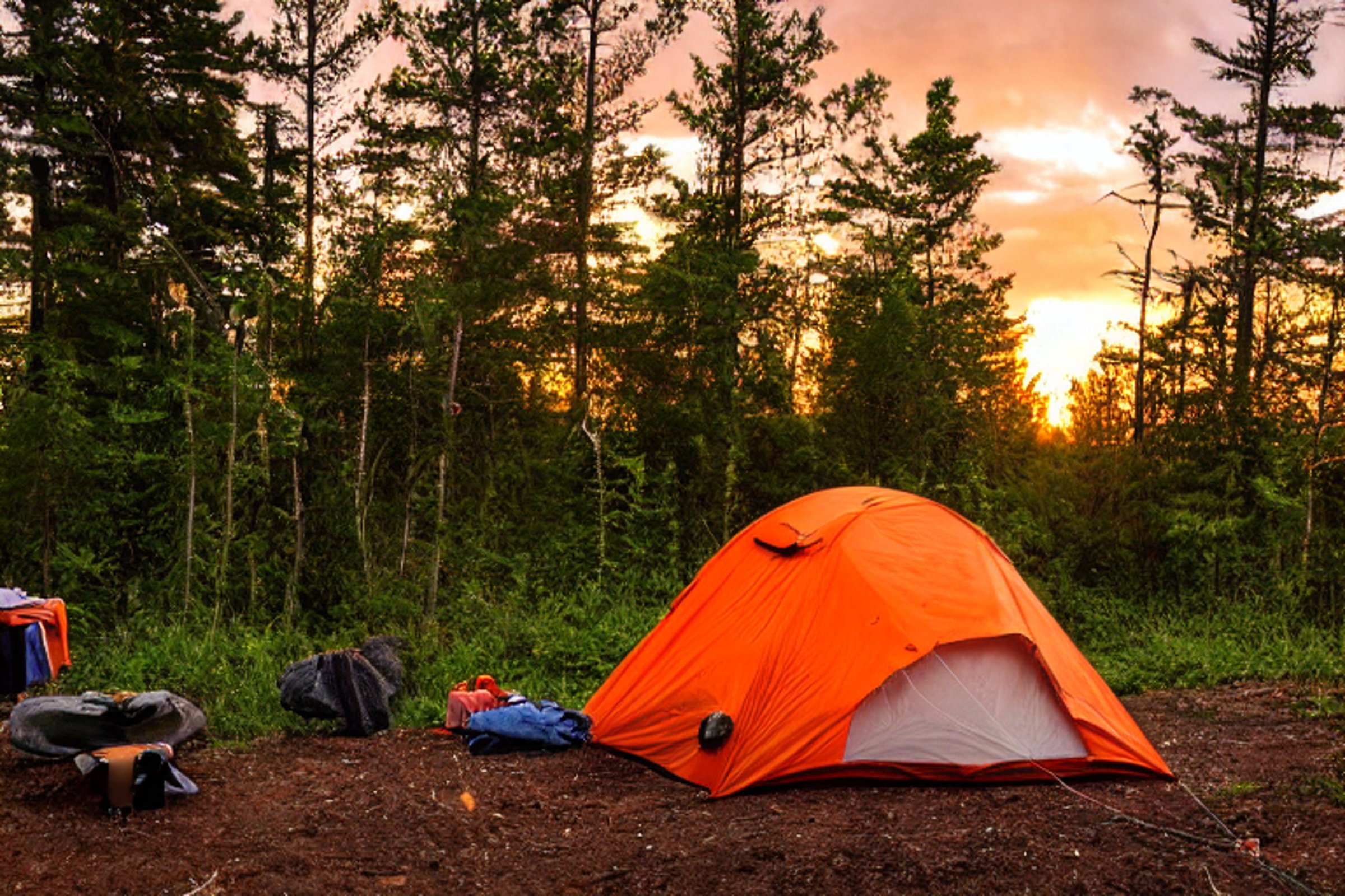 An orange tent glows beside a warm campfire on a dirt clearing in North Shore, Minnesota. Tall bushes surround the campsite as storm clouds gather over the sunset. A person with a flashlight stands nearby, while clothes hang on a line and a wooden guitar rests on the ground.