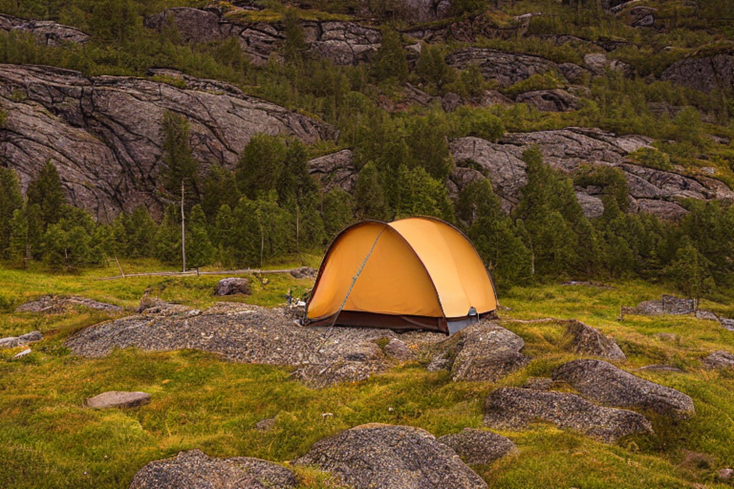A brown tent rests on a paved campsite in Norway beneath a cloudy night sky. Rocky ground and fallen leaves surround a small dirt patch, while the distant moon adds a quiet glow.