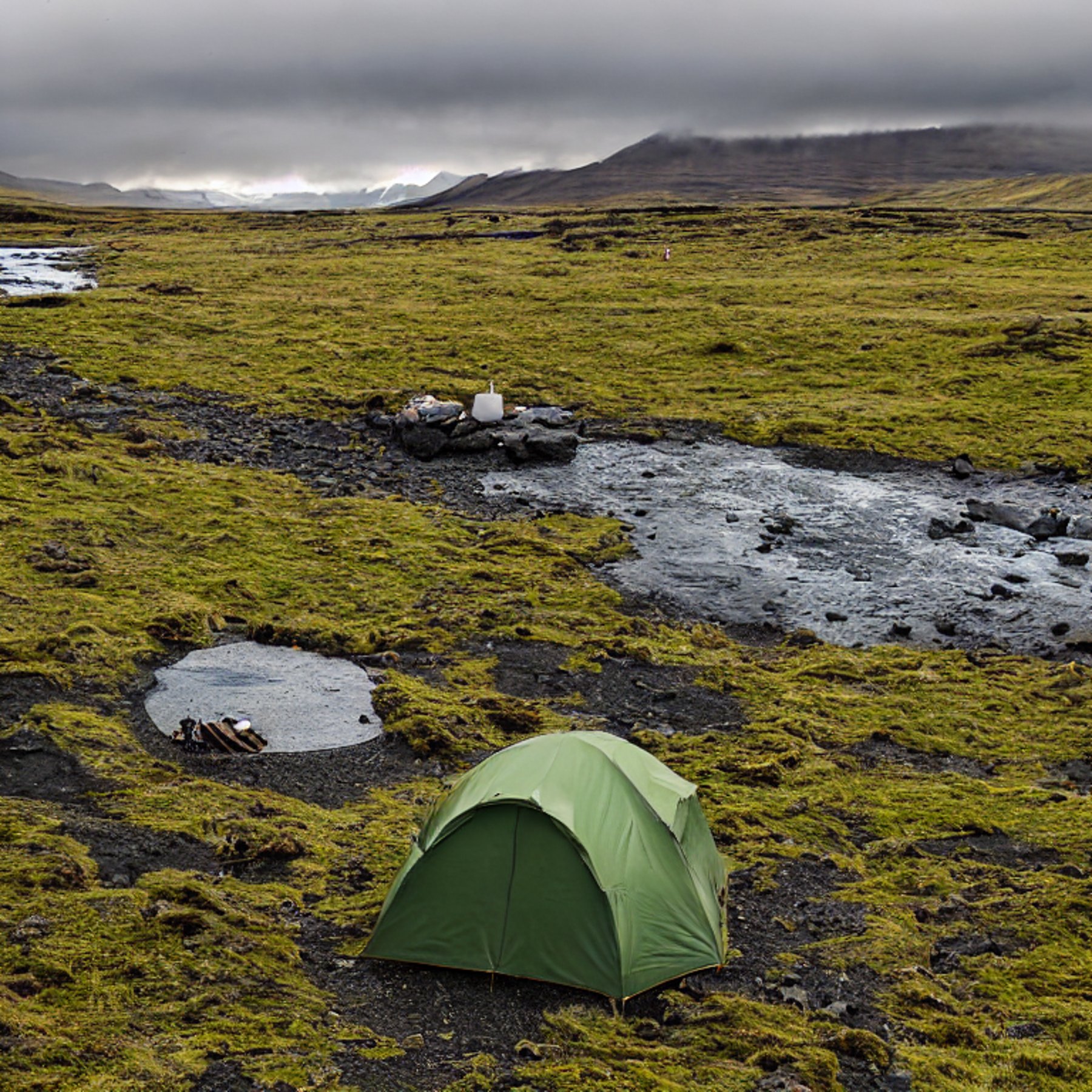 A green tent rests beside a paved campsite in Iceland, glowing in the soft light of golden hour. A clear stream winds through the camp, with a backpack and large log near the campfire. Surfers and cloudy skies add to the wild, peaceful mood.