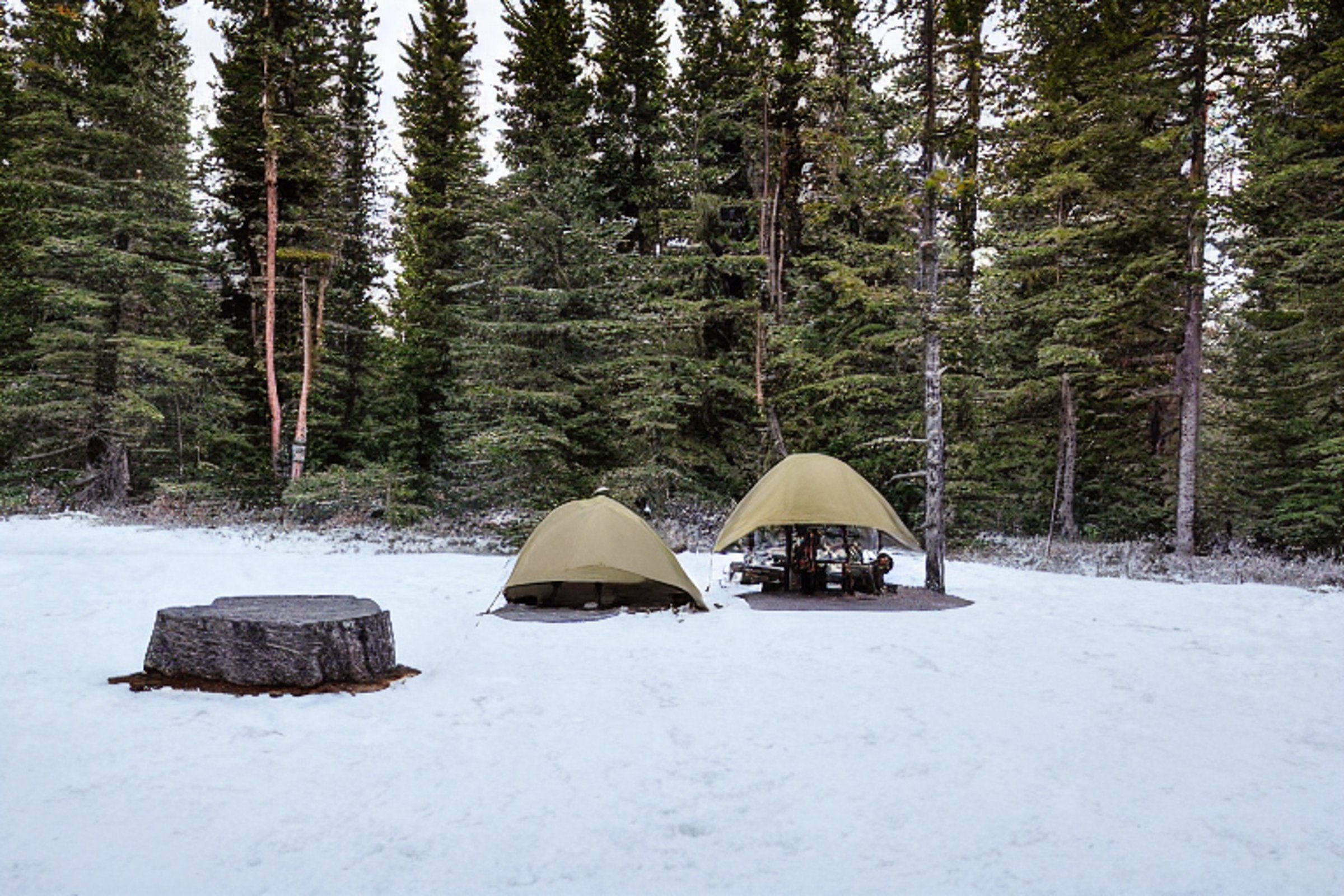 A tan tent rests on a paved campsite in snowy Banff National Park. Stone slabs and a crackling campfire create a cozy spot, while a hammock hangs between the trees. Rolling hills glow softly under the sunset sky.