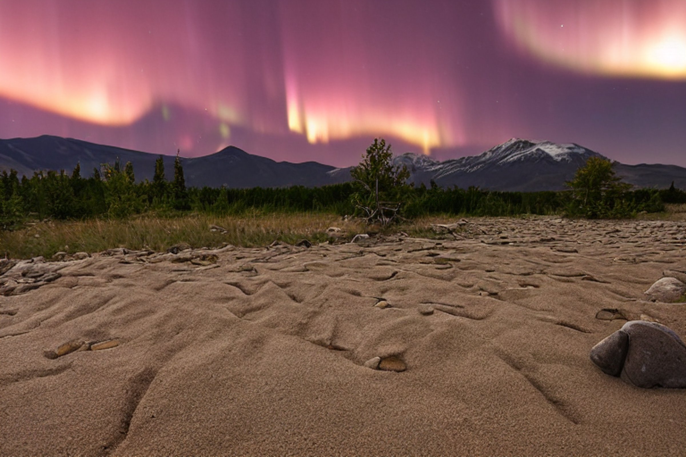 A cozy tan tent rests on a paved campsite beneath glowing green northern lights. Dark mountains and sandy dunes rise in the rainy night, with stones and a small twig scattered around the camp.