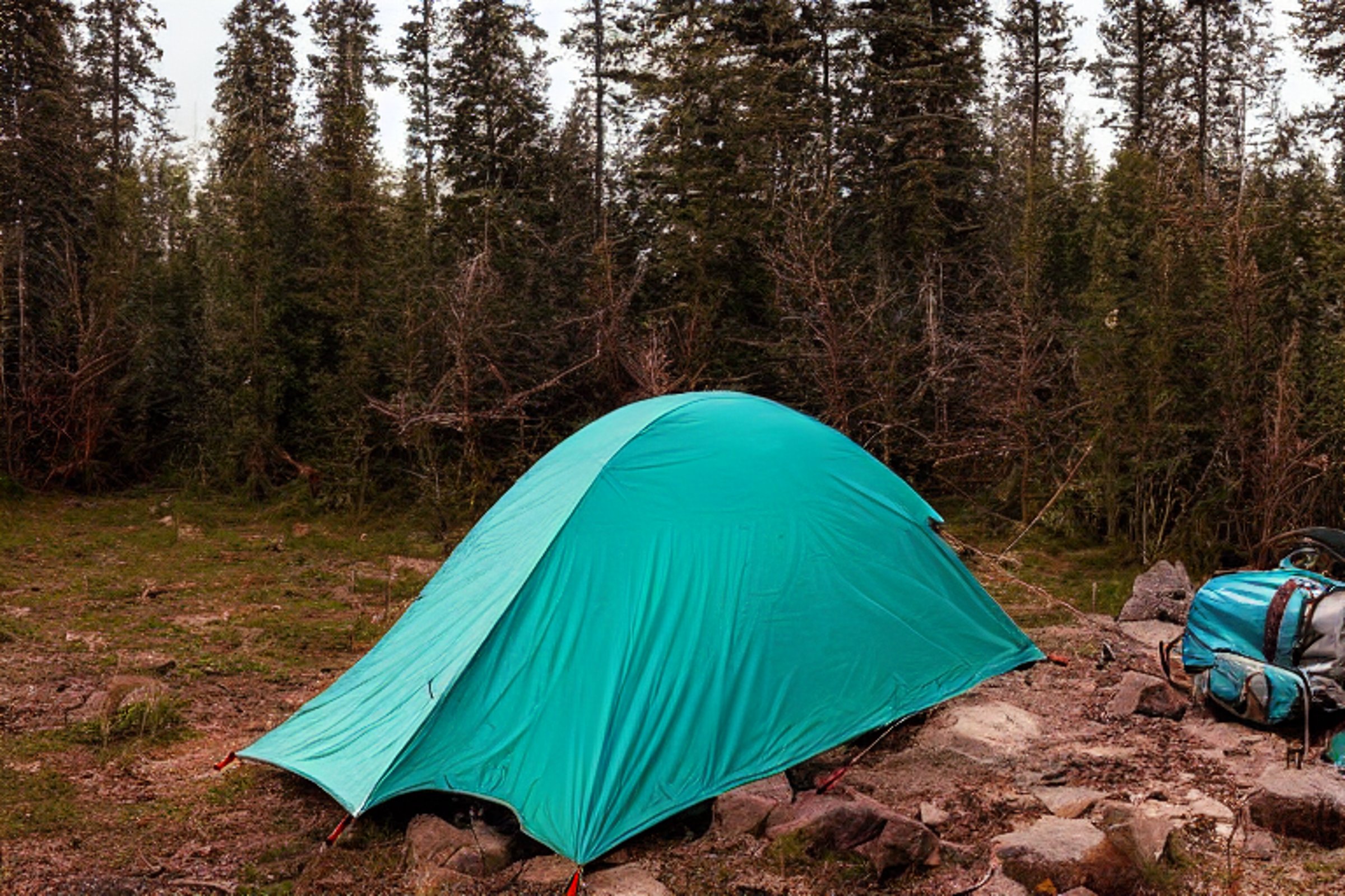 A teal tent rests on a rocky campsite beside a quiet lake. Snowy ground, rough dirt, and a small lantern create a cozy feel as the sun sets.