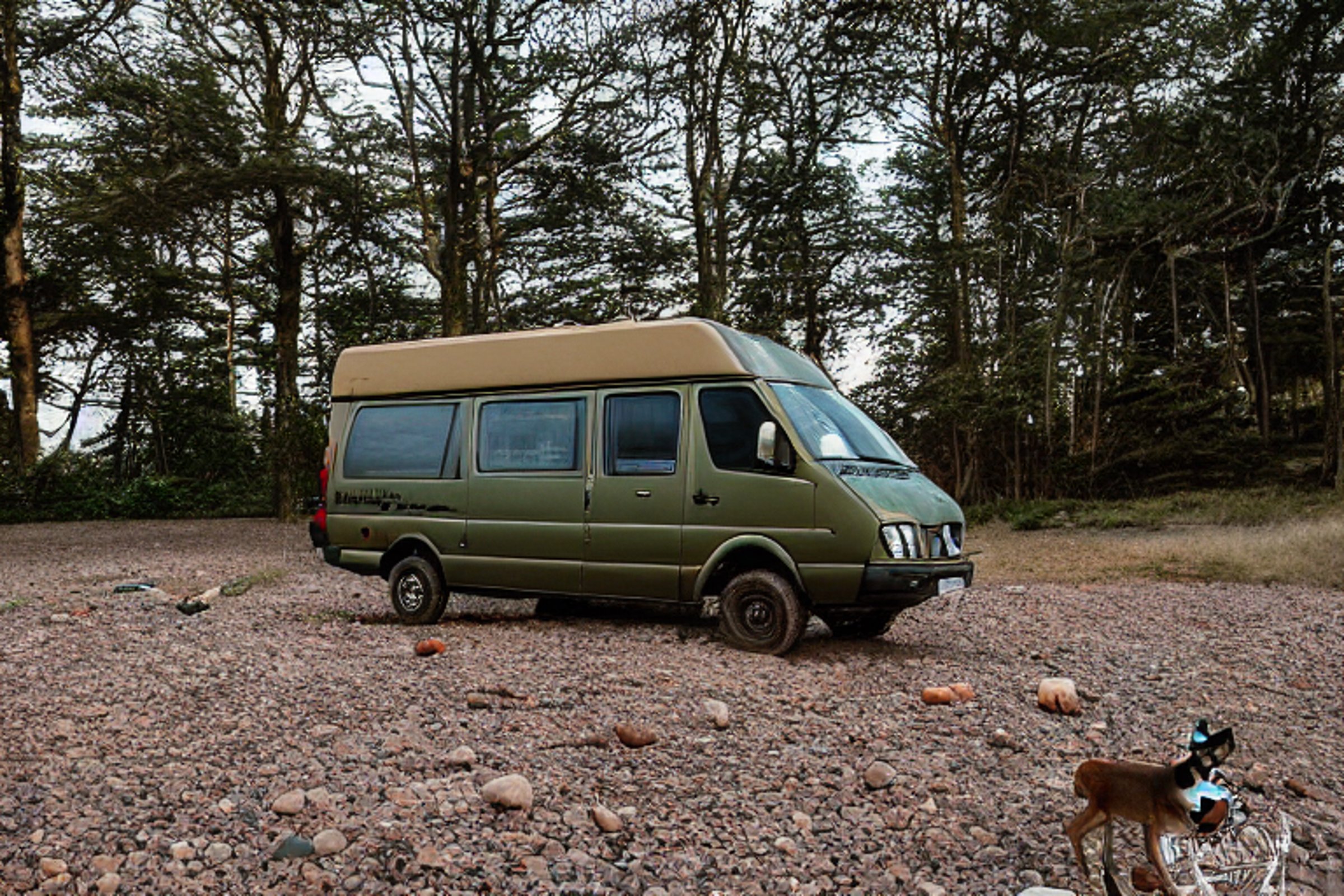 A green and tan camper van rests on a rocky patch near Cannon Beach. A warm campfire glows as the sun sets, with a deer in the distance and the moon rising above the coast.