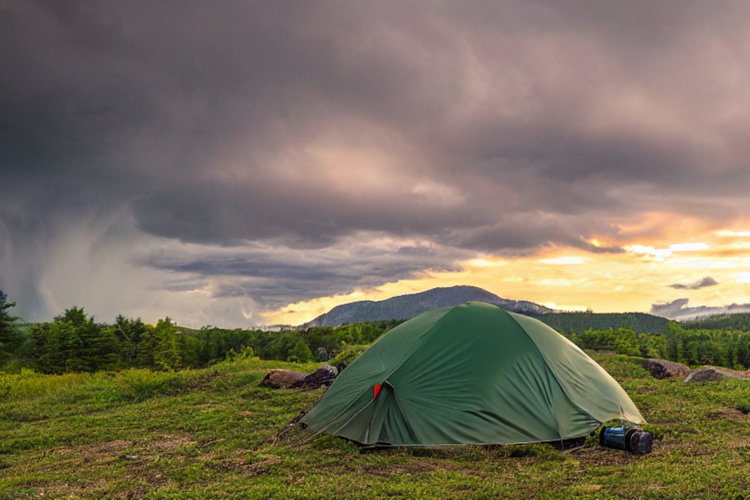 A green tent rests on a dirt campsite beside a gentle stream. Rolling hills and a mountain rise in the distance under a stormy sunset sky. A camper’s flashlight glows softly nearby.