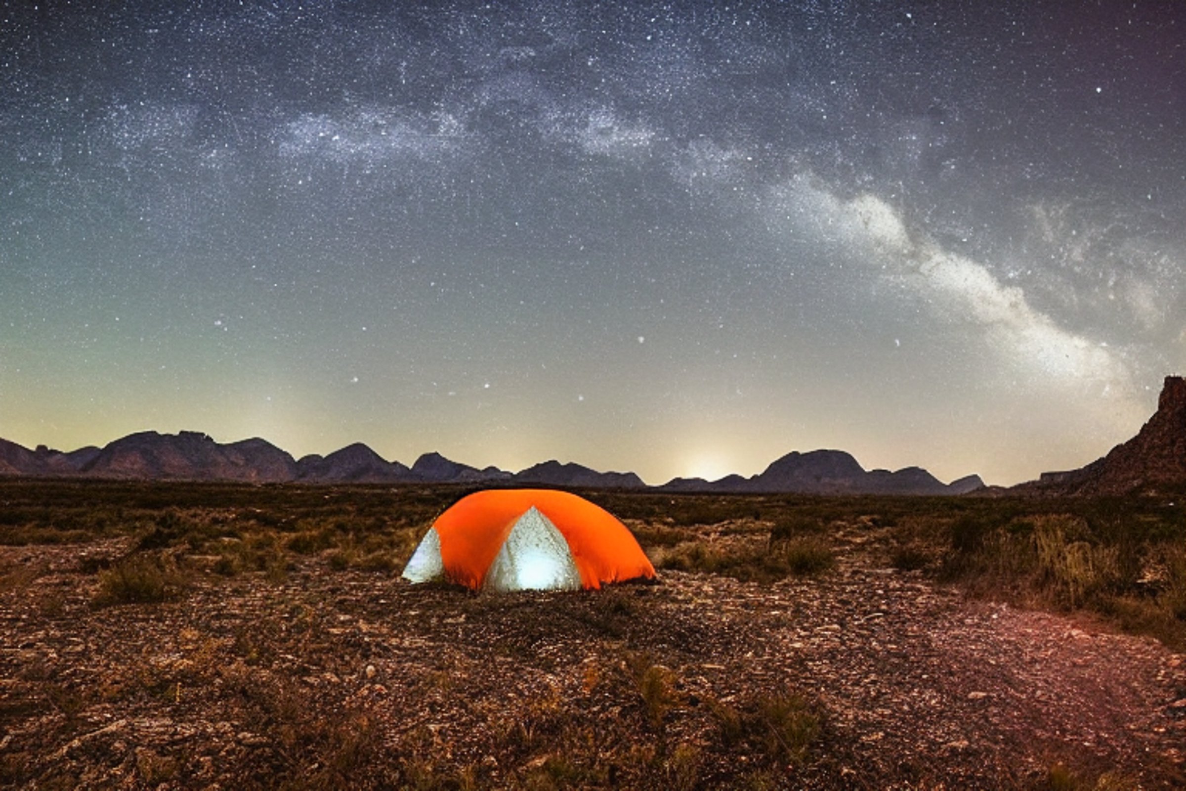 A gray and orange tent rests on a paved campsite in Big Bend National Park. Tall grass, scattered stones, and a berry-covered bush surround the muddy ground. Rainy clouds drift beneath a moonlit, star-filled sky.