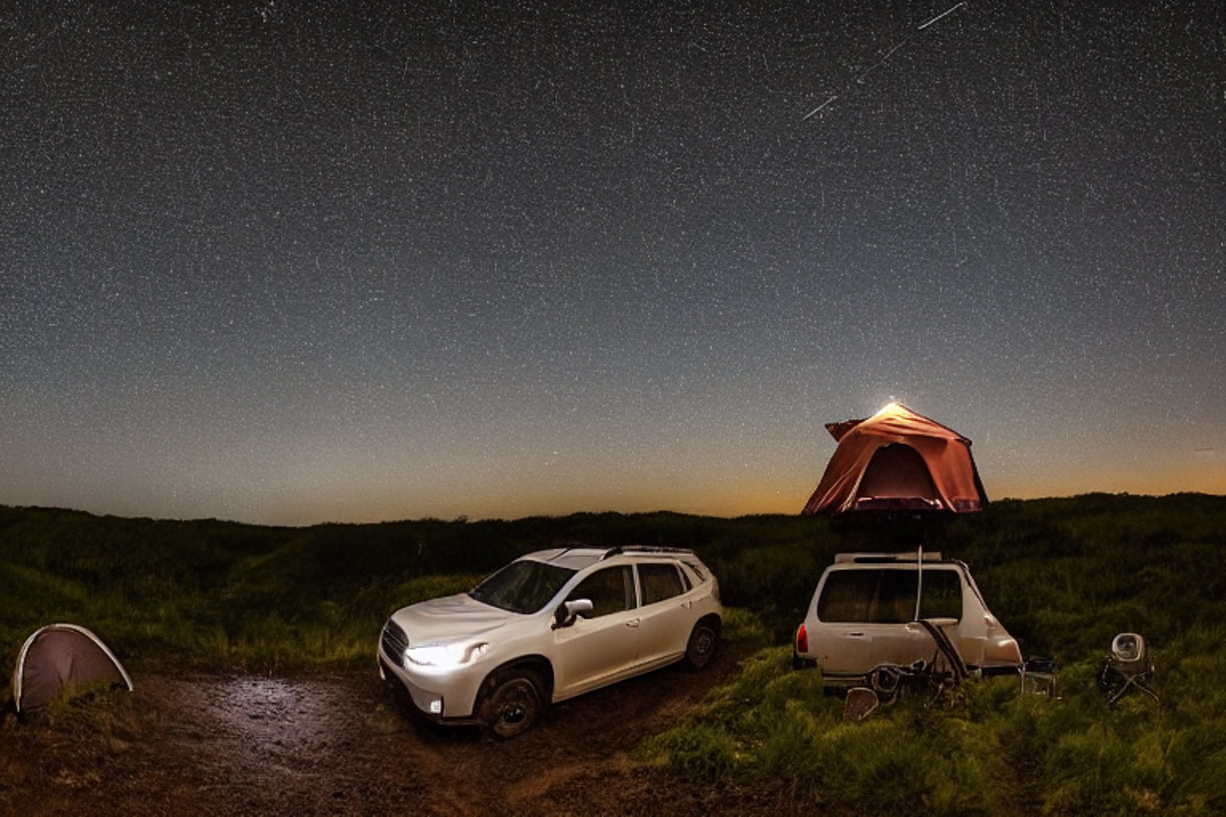 A black Subaru with a rooftop tent rests on a rough beach campsite under a star-filled, rainy sky. A small stream winds through the stones and dirt, with camping chairs nearby. The ocean adds a wild, peaceful feel to the nighttime scene.