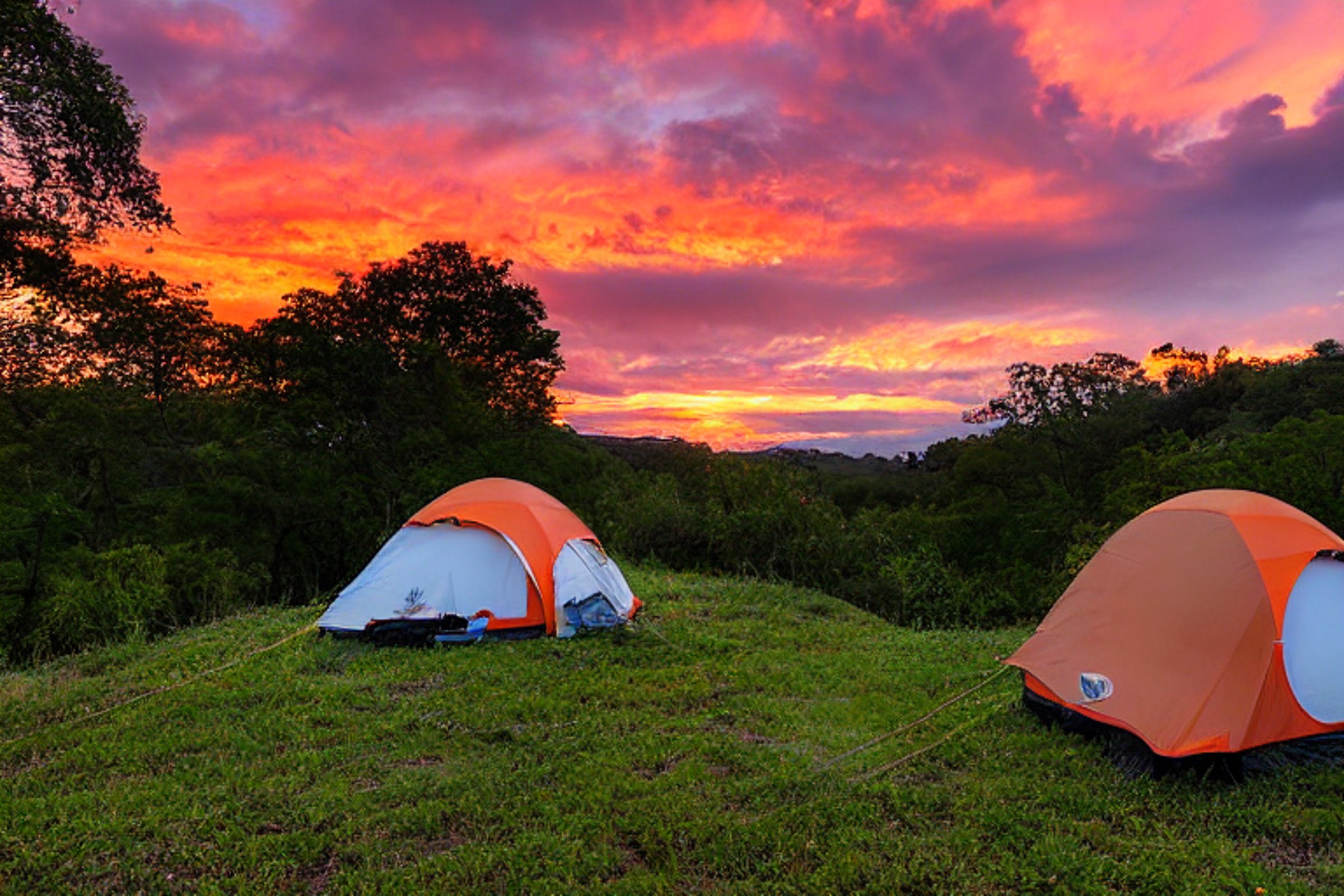 An orange tent rests on a paved campsite as rain falls over the rolling hills. Wet leaves and stone patches surround the dirt clearing, while hikers pass by under the soft sunset sky.