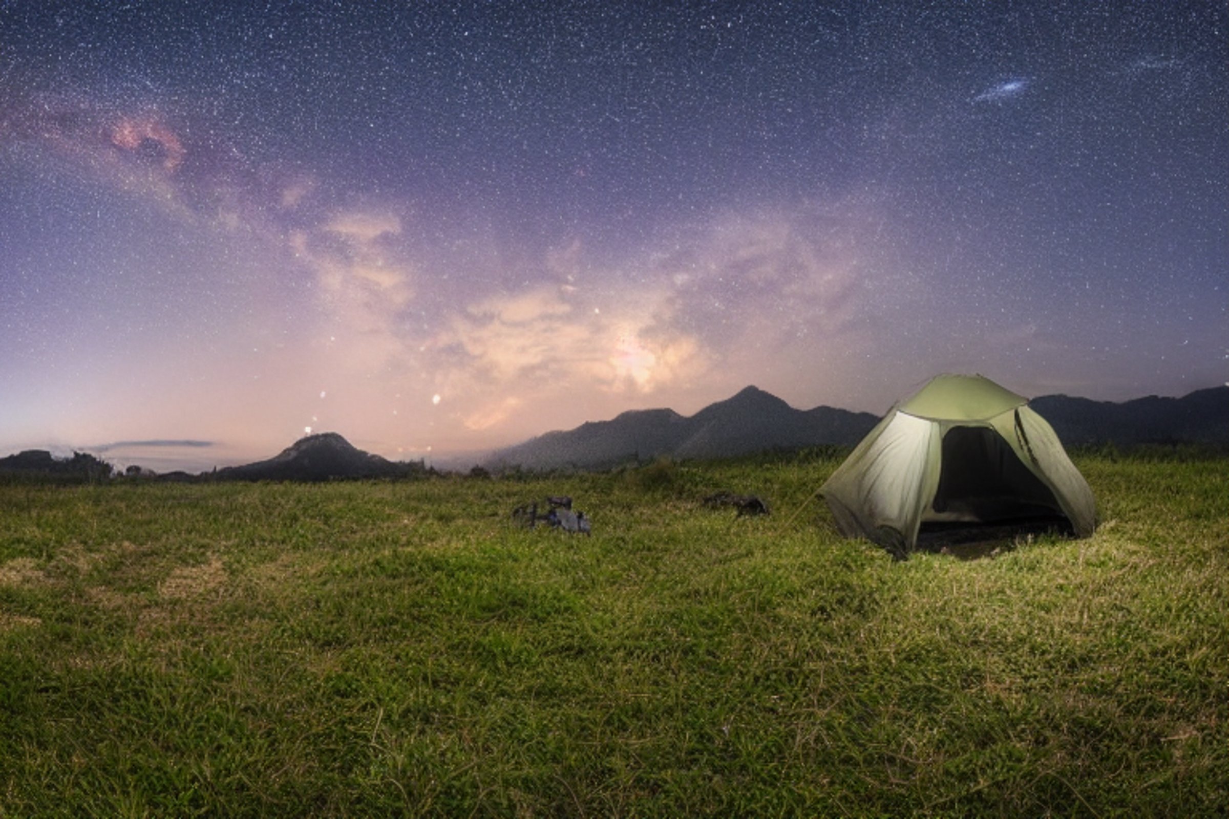 A cozy tent rests on a paved campsite beneath a star-filled, cloudy sky. Rocky ground and tall grass surround the site, with rolling hills and distant mountains adding to the peaceful night view.