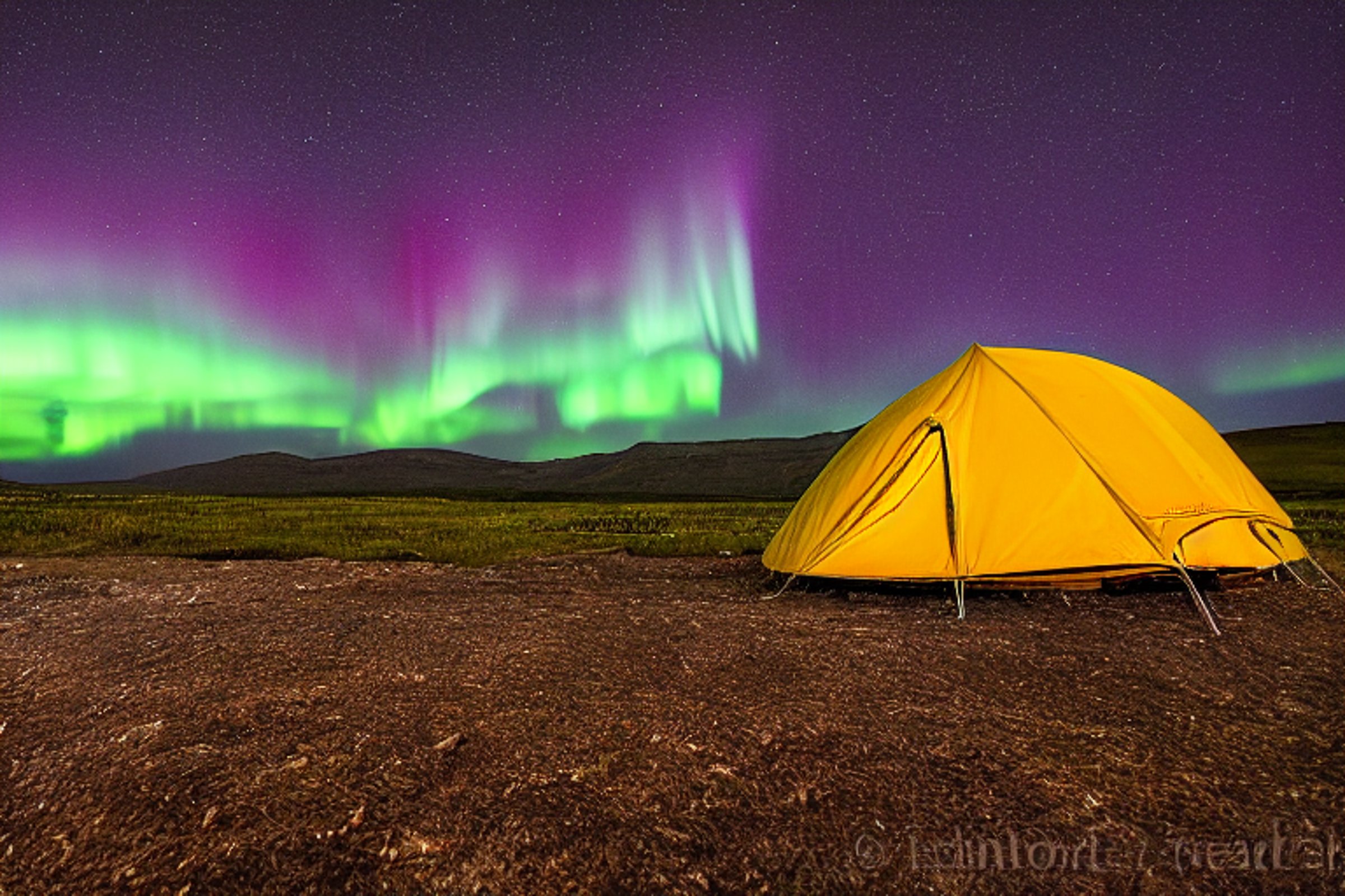 A cozy tent rests on a dirt patch beside a paved campsite in the Idaho plains. Bright northern lights dance across the stormy night sky, with a distant moon glowing above. Small flowers add a gentle touch to the rugged ground.