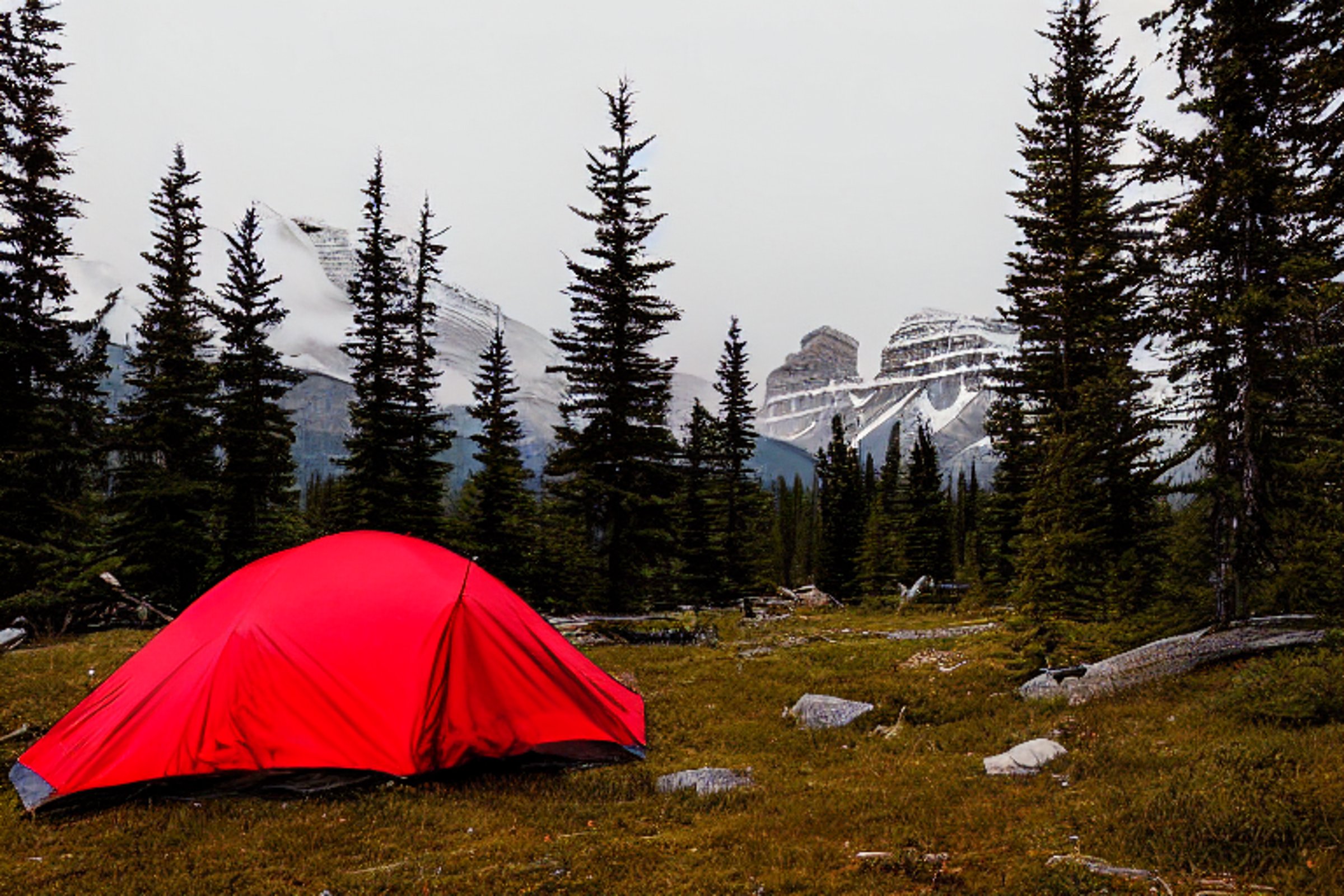 A red and white tent rests on a quiet dirt campsite in Banff National Park. Tall trees, misty air, and mountains create a calm dusk setting, while a distant camper’s flashlight glows softly.