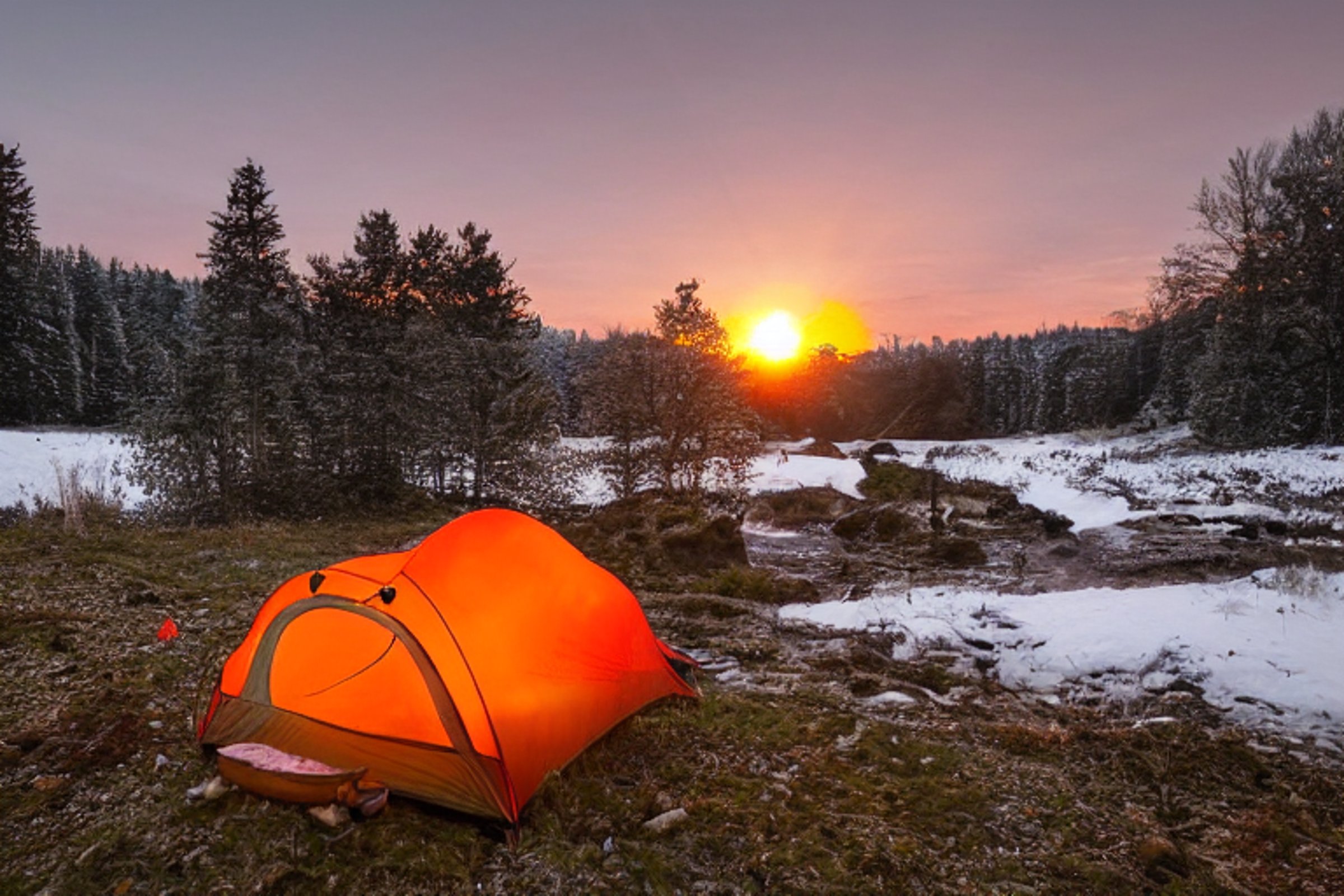 An orange tent sits beside a warm campfire near a quiet river. Snowy ground, smooth stones, and fallen leaves surround the campsite, while a deer stands far away beneath the glowing sunset.