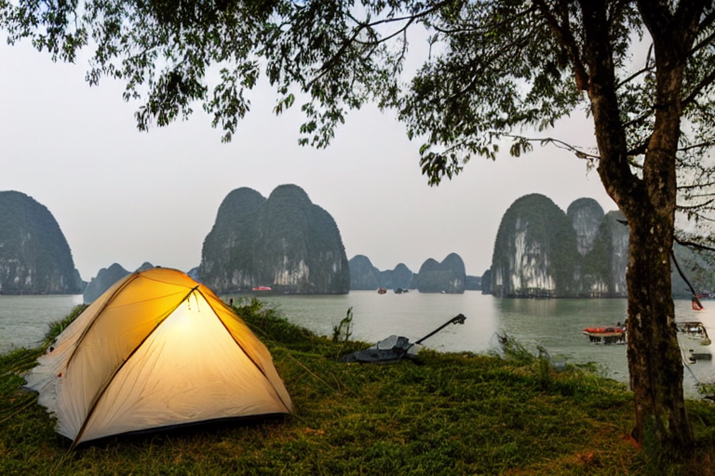 A blue tent rests on a paved campsite in Halong Bay, surrounded by tall trees and misty mountains. Warm golden light, a glowing lantern, and clothes drying on a line create a cozy evening mood.