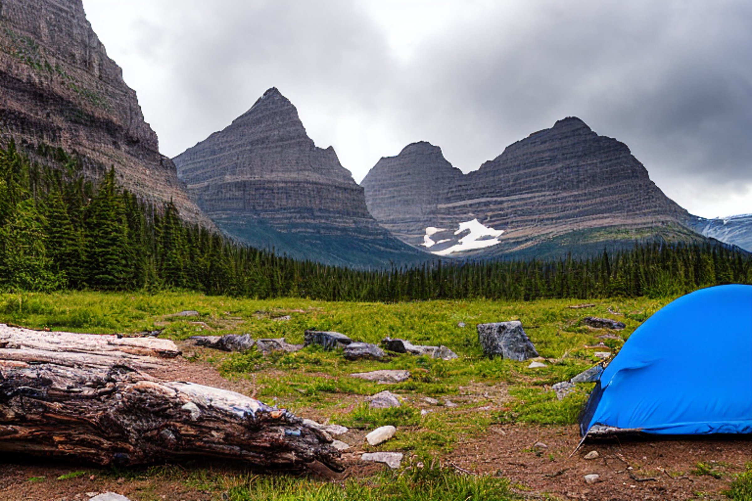 A blue and gray tent rests on a dirt patch at a paved campsite in Glacier National Park. Large rocks, boulders, and a fallen log surround the campfire area as hikers pass nearby. Stormy dusk clouds create a cool, wild atmosphere.