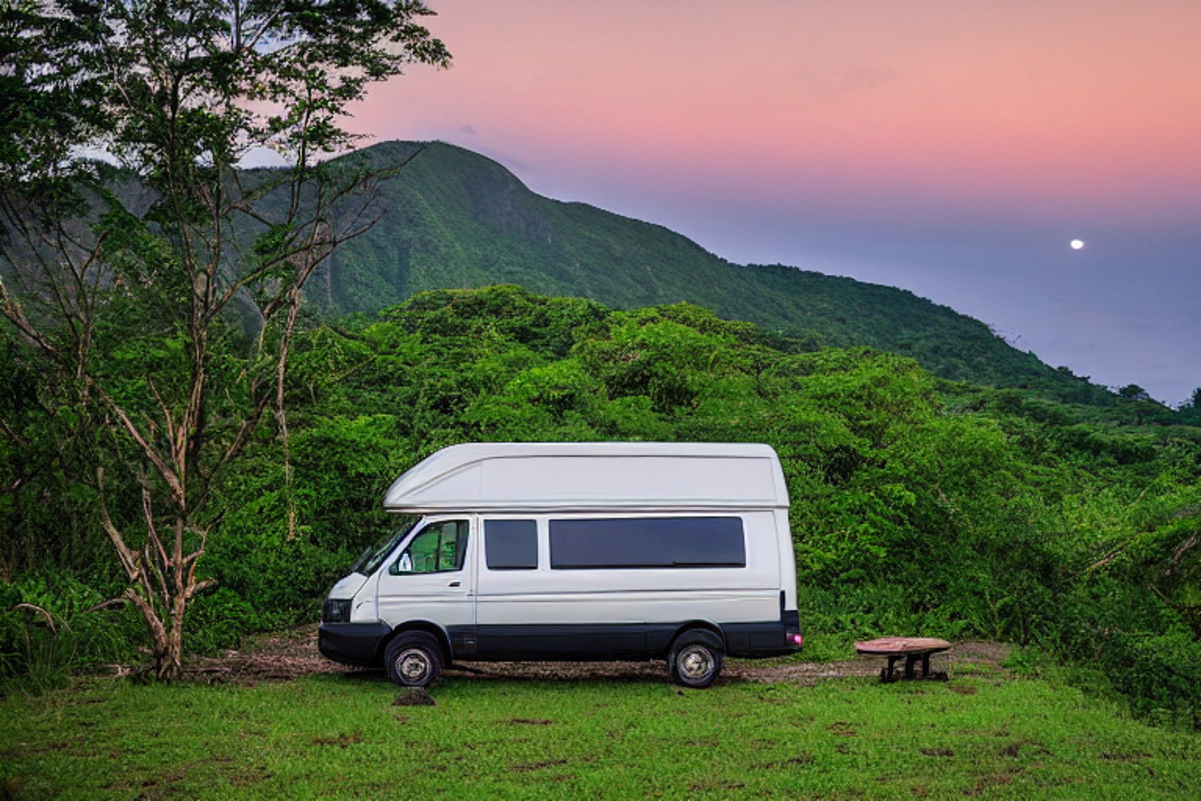 A forest-green van rests on a quiet dirt campsite in Soufrière, St. Lucia. Rolling hills, small trees, and a large rock surround the camp, while a pale moon glows through the snowy dusk.