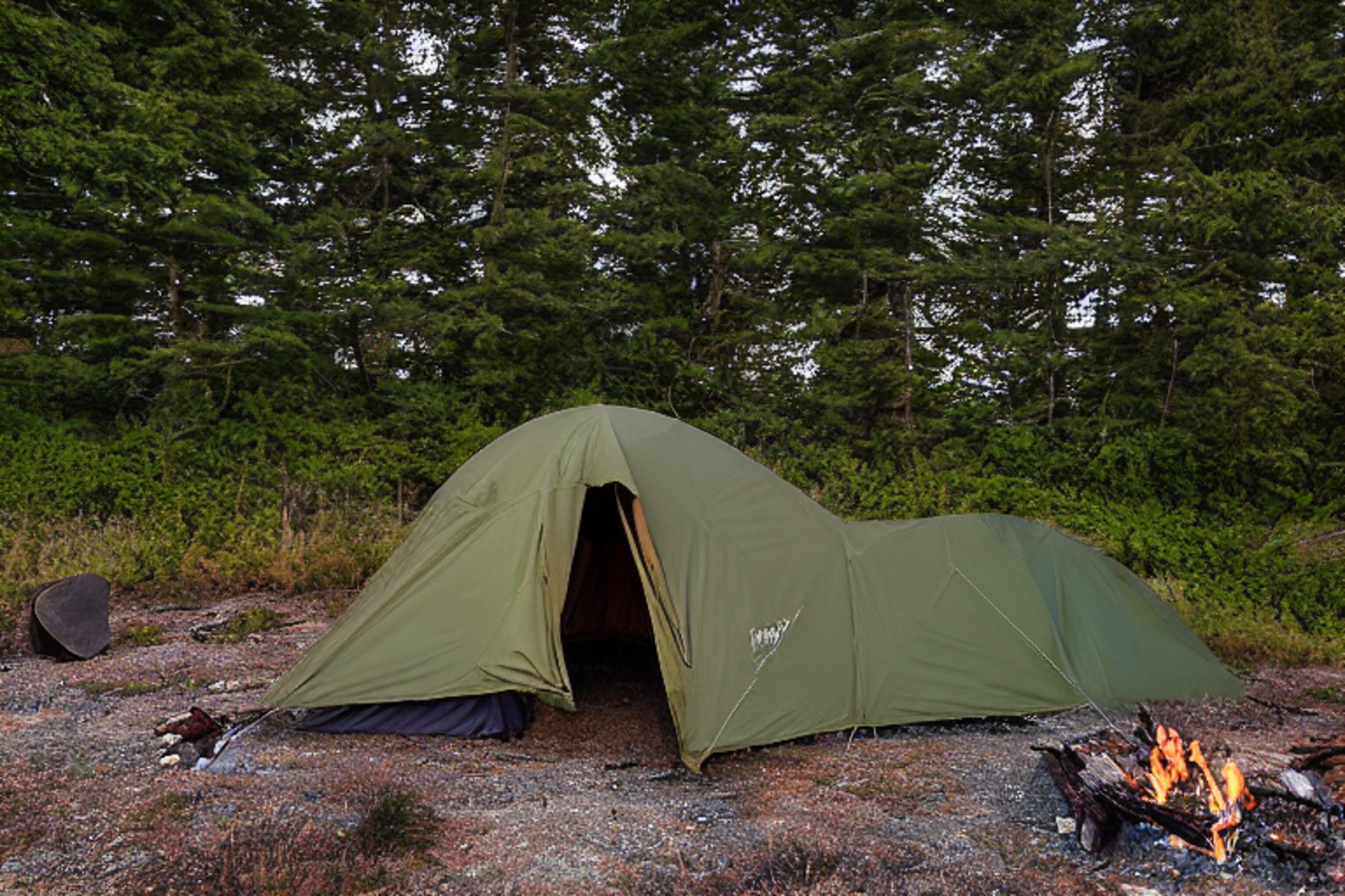 A green and tan tent rests on a paved campsite along the coast of Maine. Bushes, a large rock, and a wood log surround the dirt campfire area as clouds drift across the dusky sky. A pale moon glows in the distance, creating a calm evening mood.