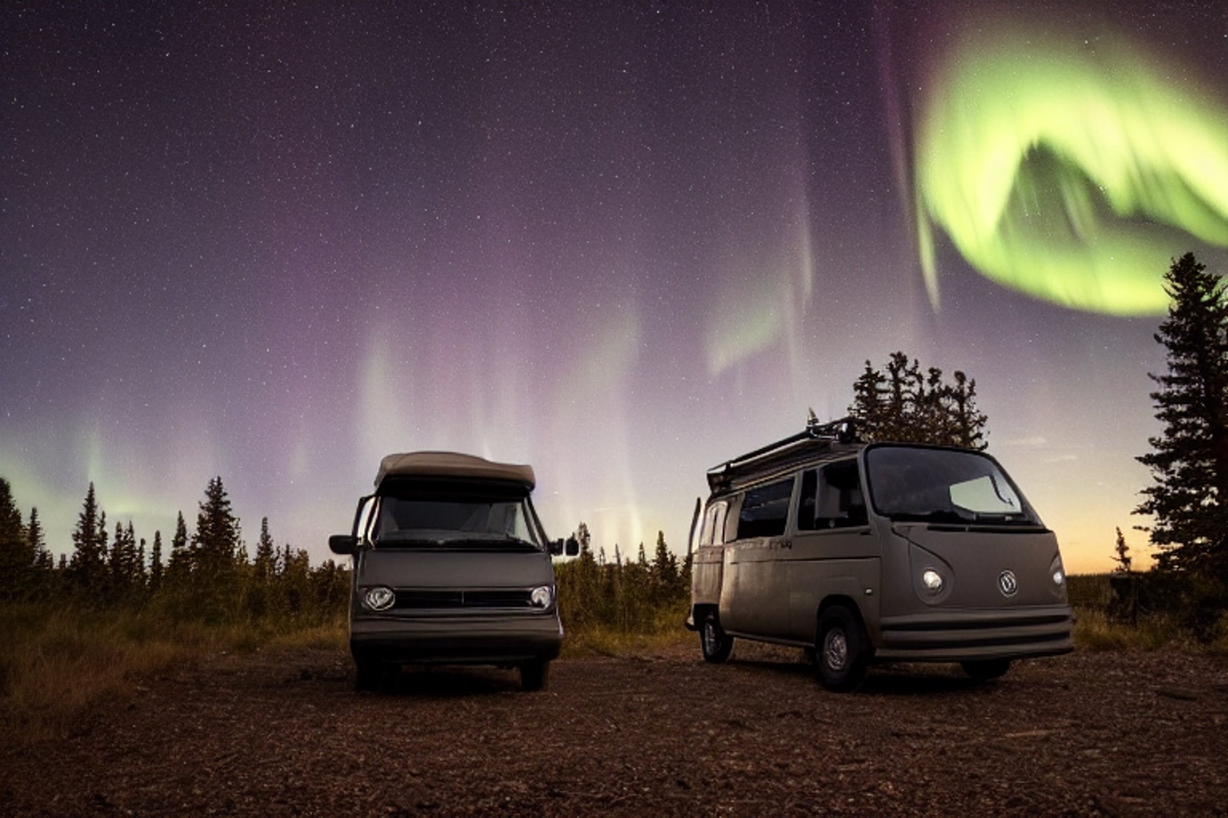 A black camper van rests on a rough dirt campsite beneath the glowing northern lights. The Grand Canyon stretches into the dark distance, while a lantern shines nearby and a lone camper walks with a flashlight.