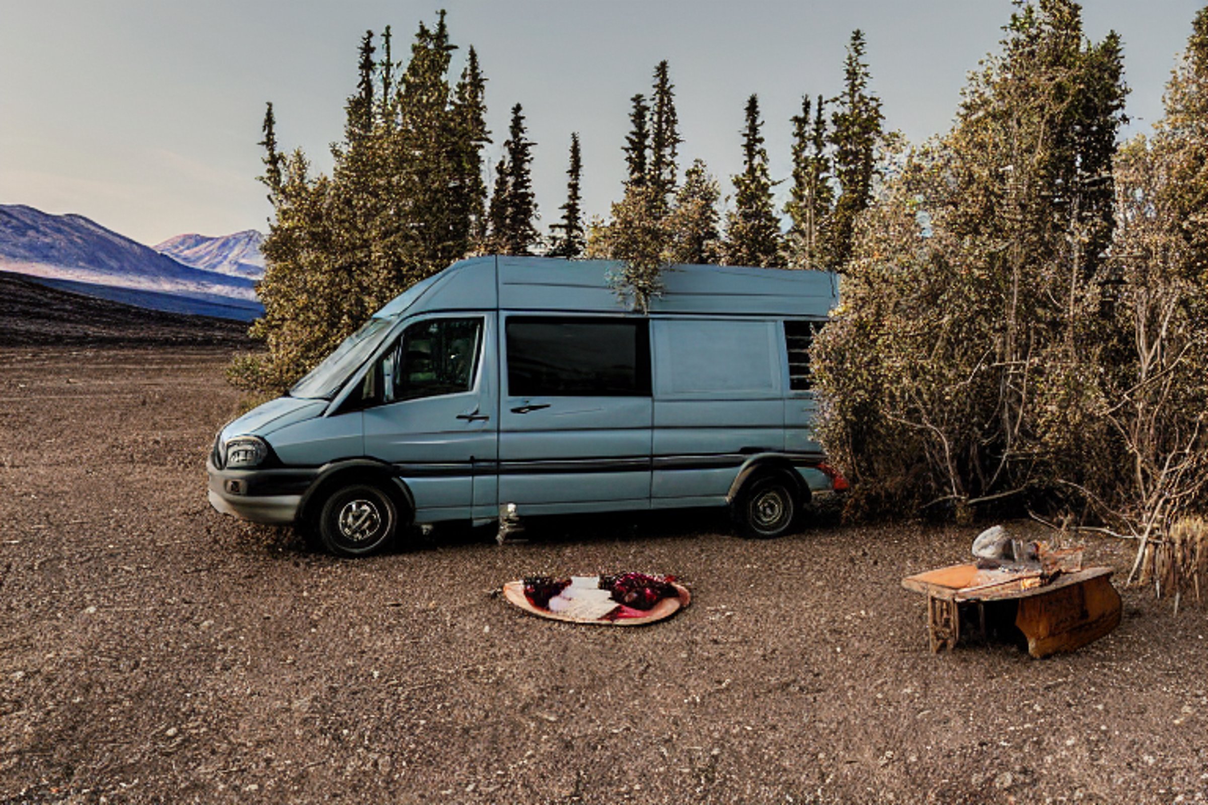 A cozy camper van rests on a rough dirt patch beneath glowing northern lights. Small flowers, berry bushes, and a wooden guitar add charm to the foggy desert campsite.