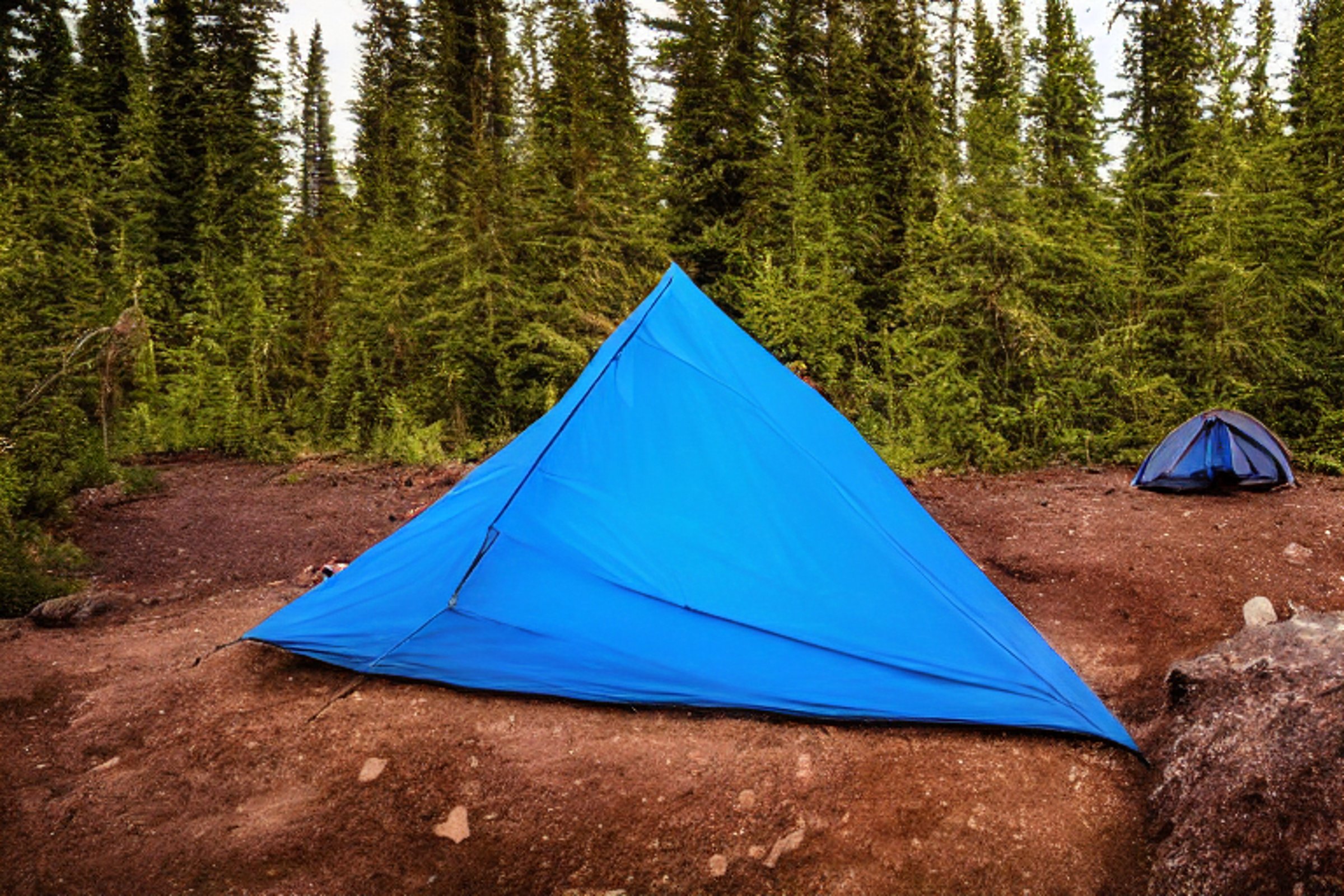 A blue tent rests on a quiet dirt campsite beside a sparkling creek. Large bushes and stone patches surround the area, while colorful northern lights glow overhead. In the distance, a camper’s flashlight shines through the dark.