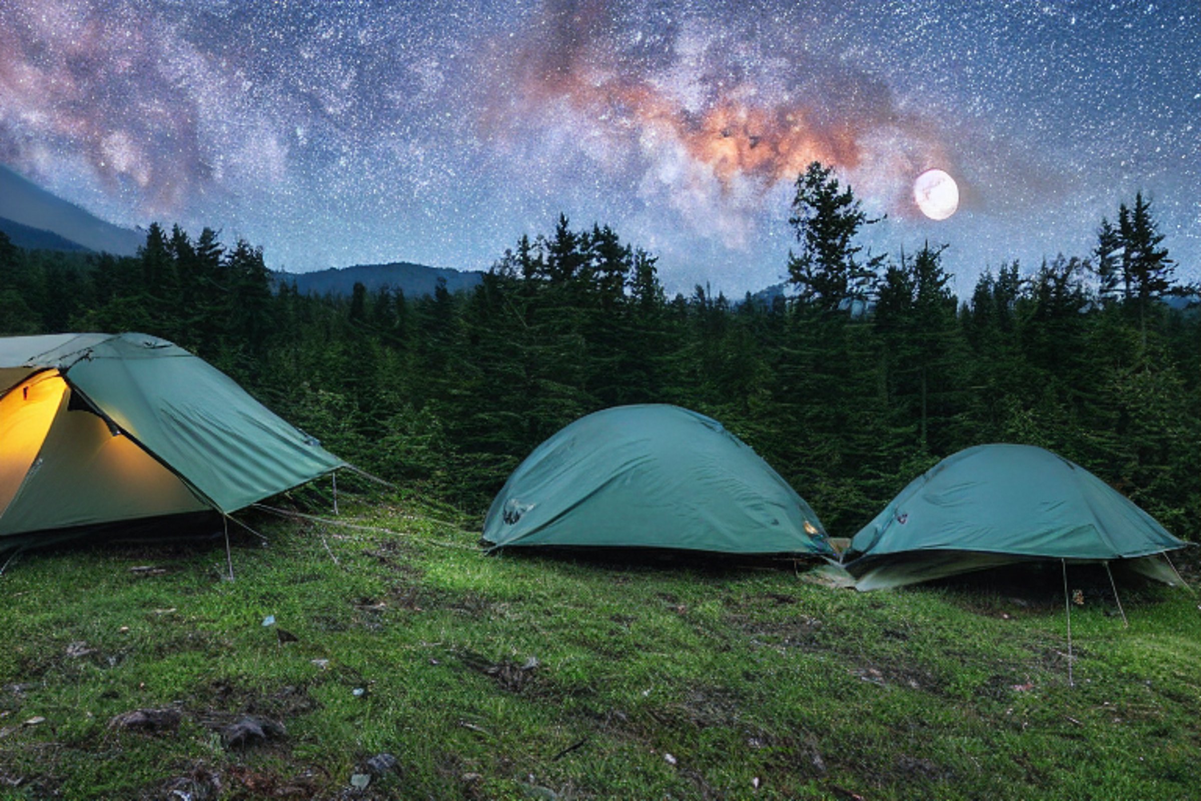 A forest green tent rests on a rocky dirt clearing in the Smoky Mountains. Tall trees surround the campsite, while a soft dusk sky reveals the distant moon. A box of camping gear sits nearby, ready for adventure.