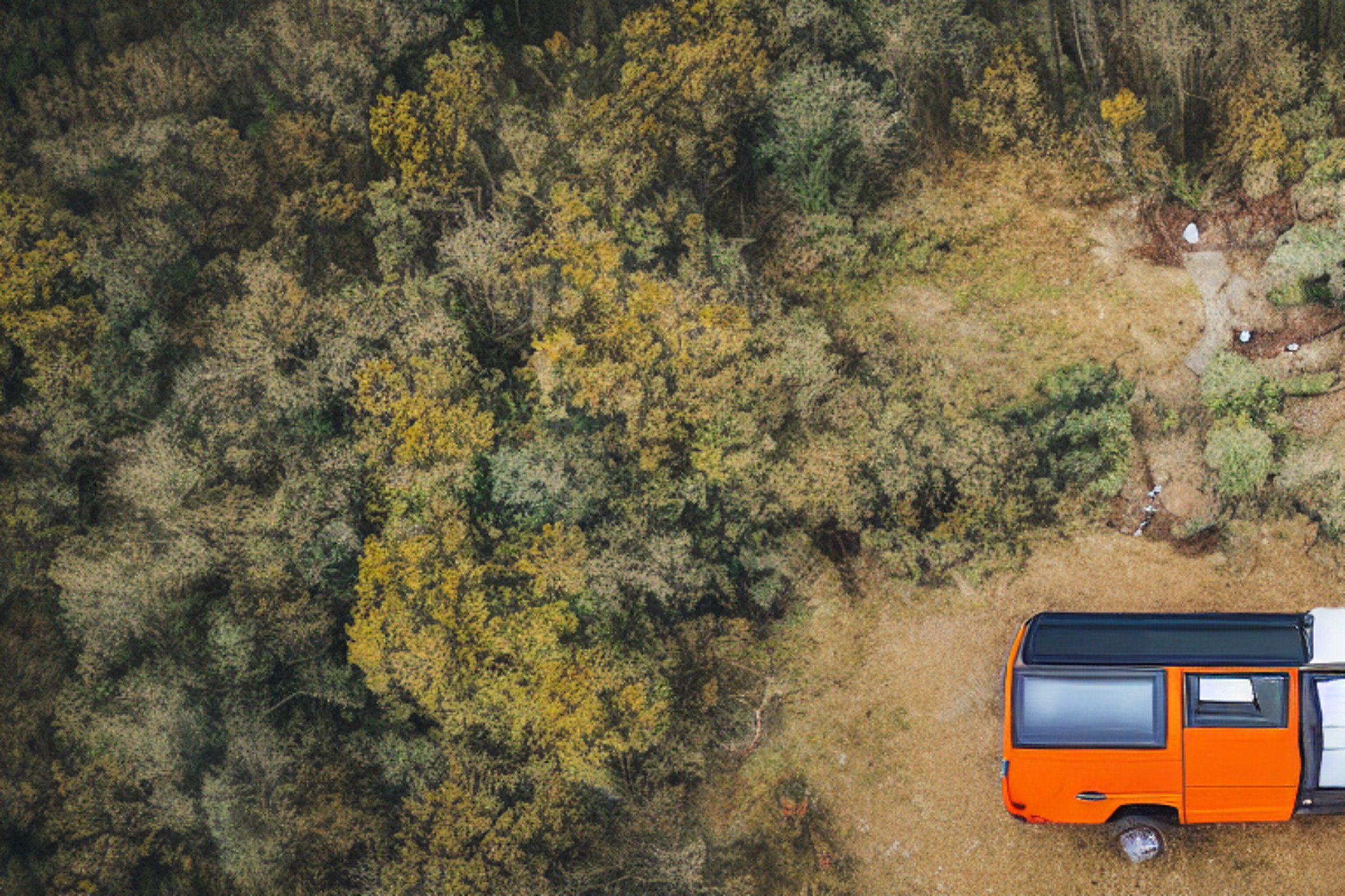 A gray and orange camper van rests on a quiet dirt campsite at sunrise. Bushes and rough land surround a winding hiking path, with a hot spring nearby. A large log sits beside the cool, empty campfire.