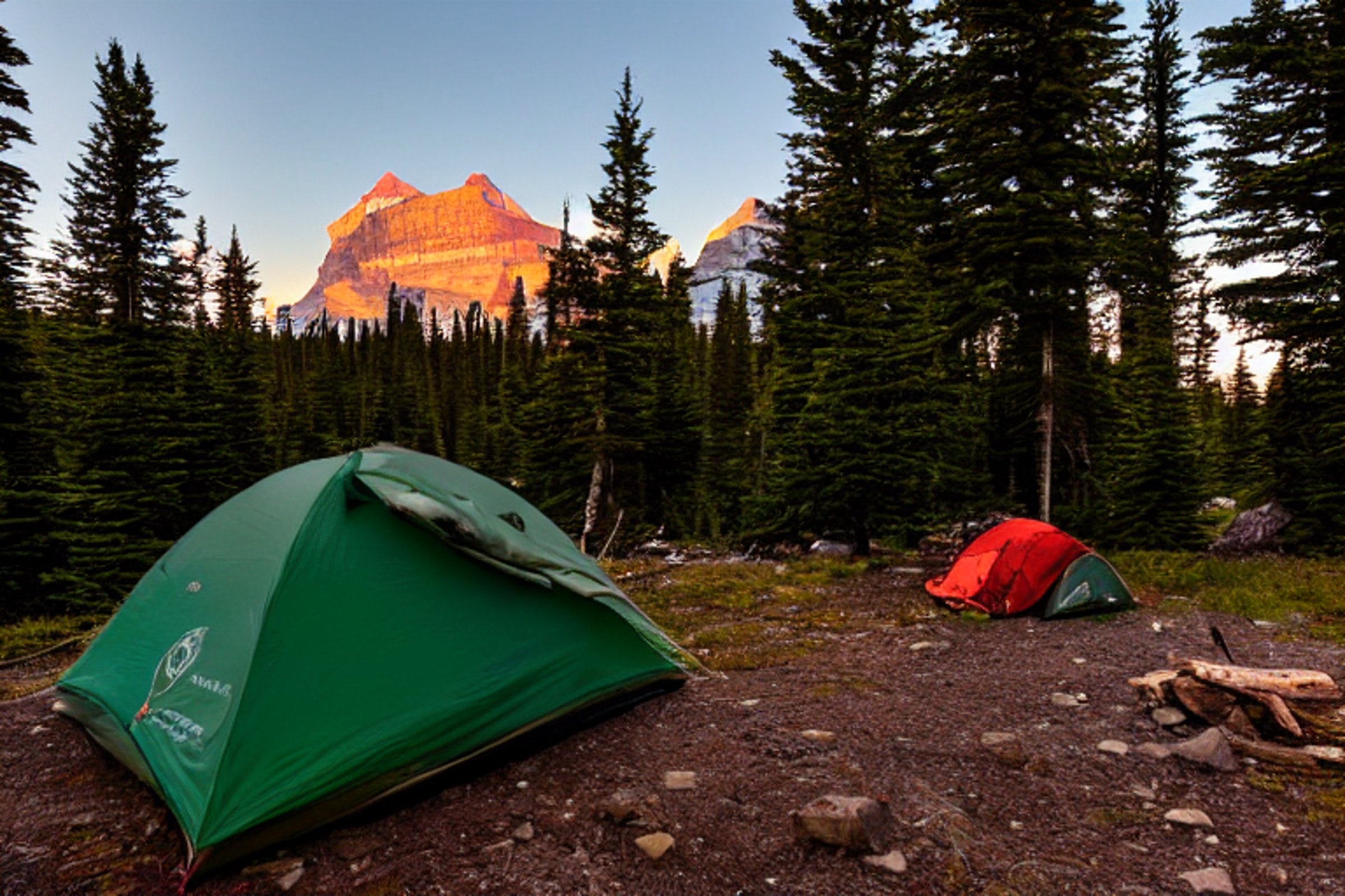 A green tent rests beside a warm campfire on a quiet dirt clearing in Glacier National Park. Tall trees surround the campsite, while bright mountains rise in the distance beneath a clear sunset sky. A backpack and small sticks lie nearby, adding to the peaceful outdoor scene.